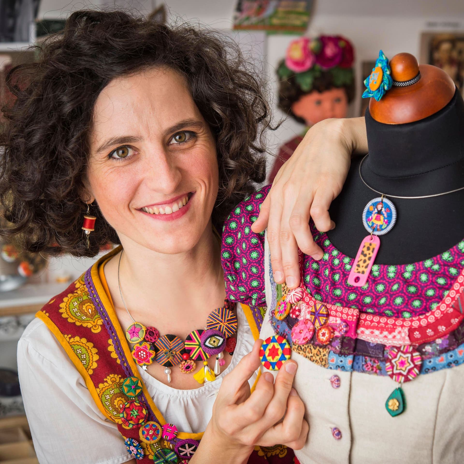 Junge Frau lächelt in die Kamera und hält einen Knopf an eine Schaufensterpuppe mit buntem Dirndl mit Posamentenknopf Collier in einem Atelier.