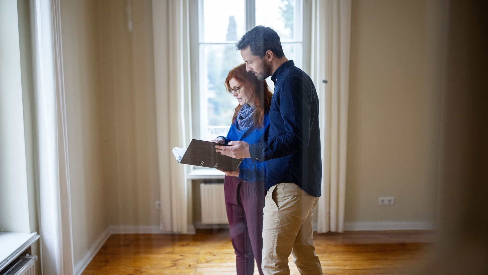 Ein Mann und eine Frau stehen in einem leeren Zimmer mit Parkettboden und halten prüfend Dokumente in den Händen.