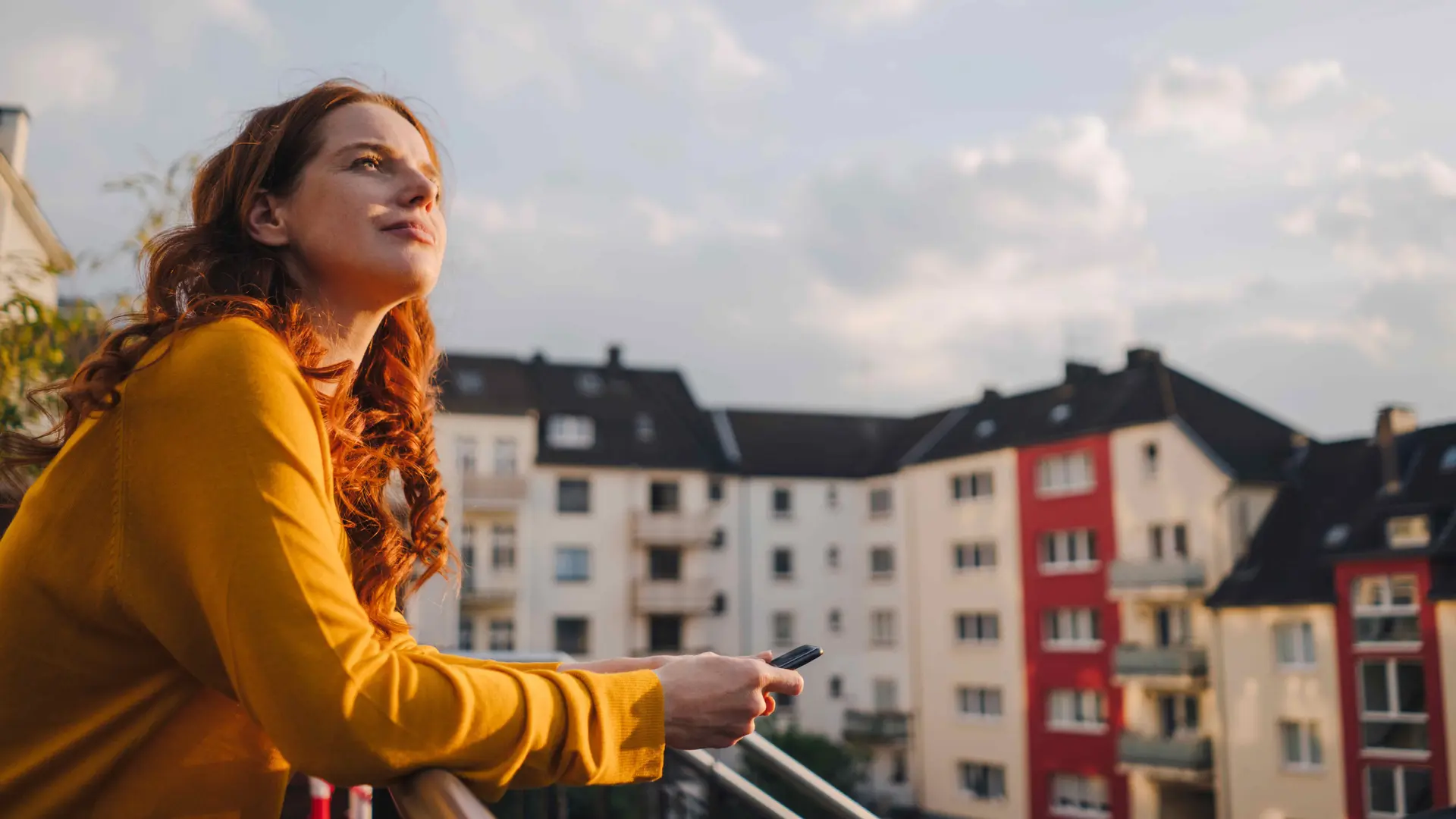 Junge Frau mit langen roten Locken steht auf einem Dachbalkon und schaut in den blauen Himmel. Im Hintergrund sind Mietswohnungen zu sehen.