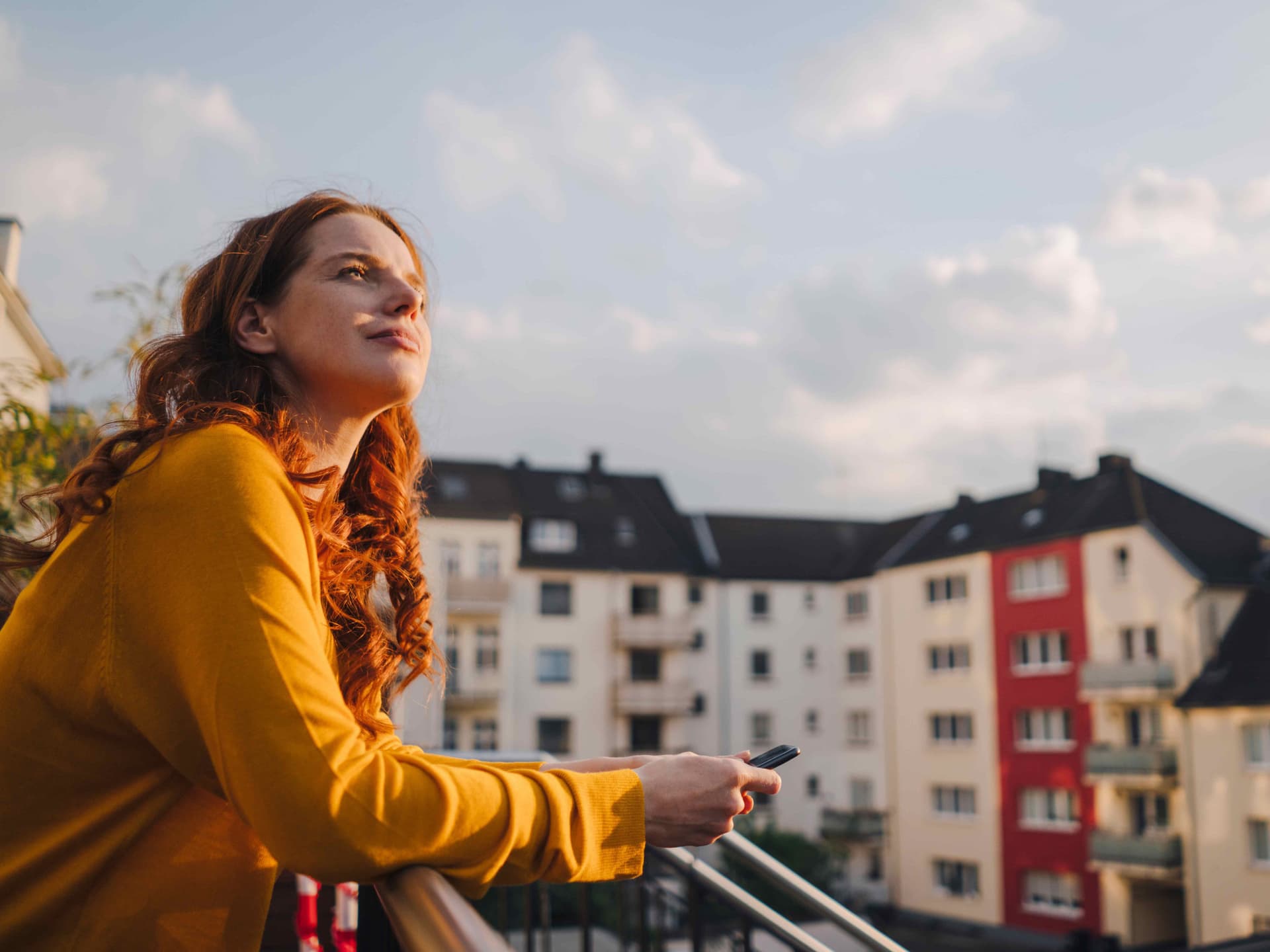 Junge Frau mit langen roten Locken steht auf einem Dachbalkon und schaut in den blauen Himmel. Im Hintergrund sind Mietswohnungen zu sehen.