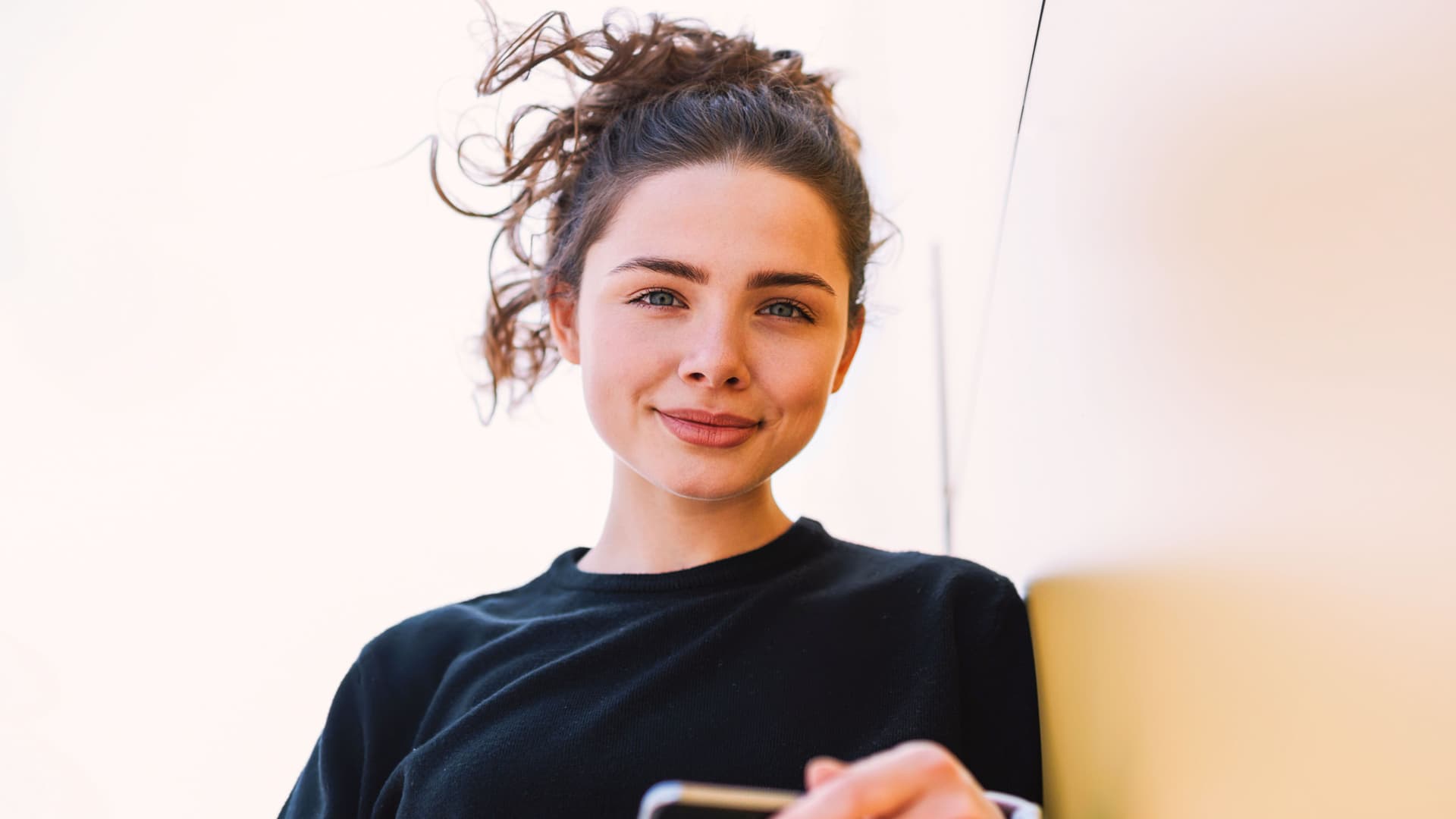 Junge Frau lehnt mit Handy an einer Wand und schaut in die Kamera.