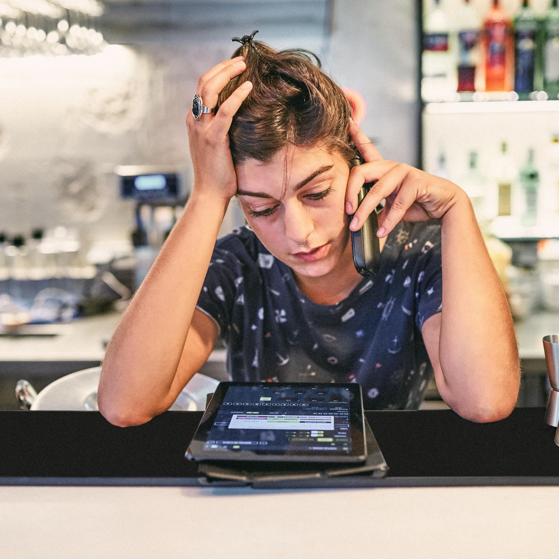 Frau hinter einer Theke schaut verzweifelt auf ein Tablet und telefoniert. Sie hat den Kopf auf eine Hand gestützt.