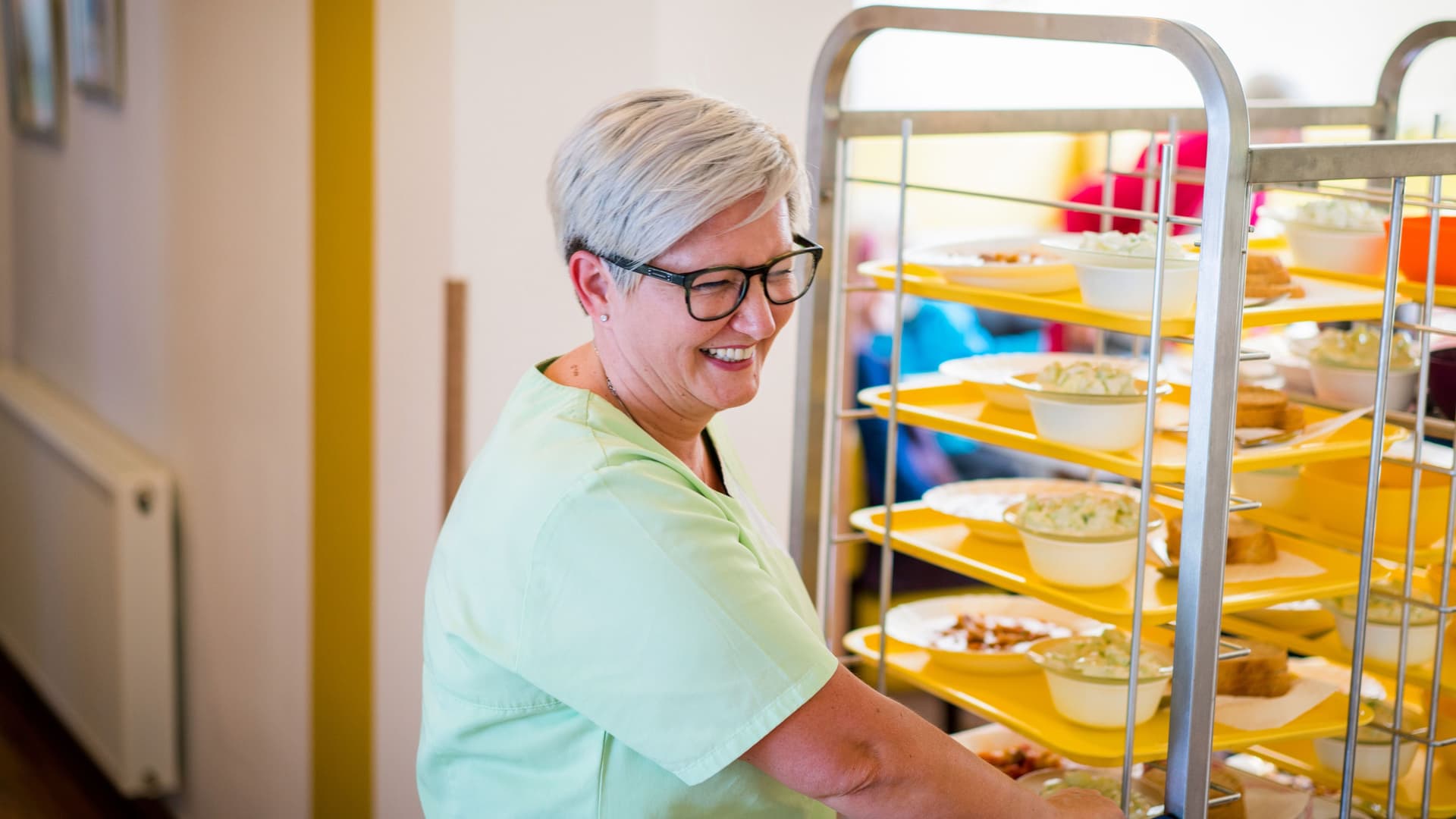 Frau in hellgrünem Kittel schiebt einen Wagen mit Essenstabletts durch eine Tür und lächelt.