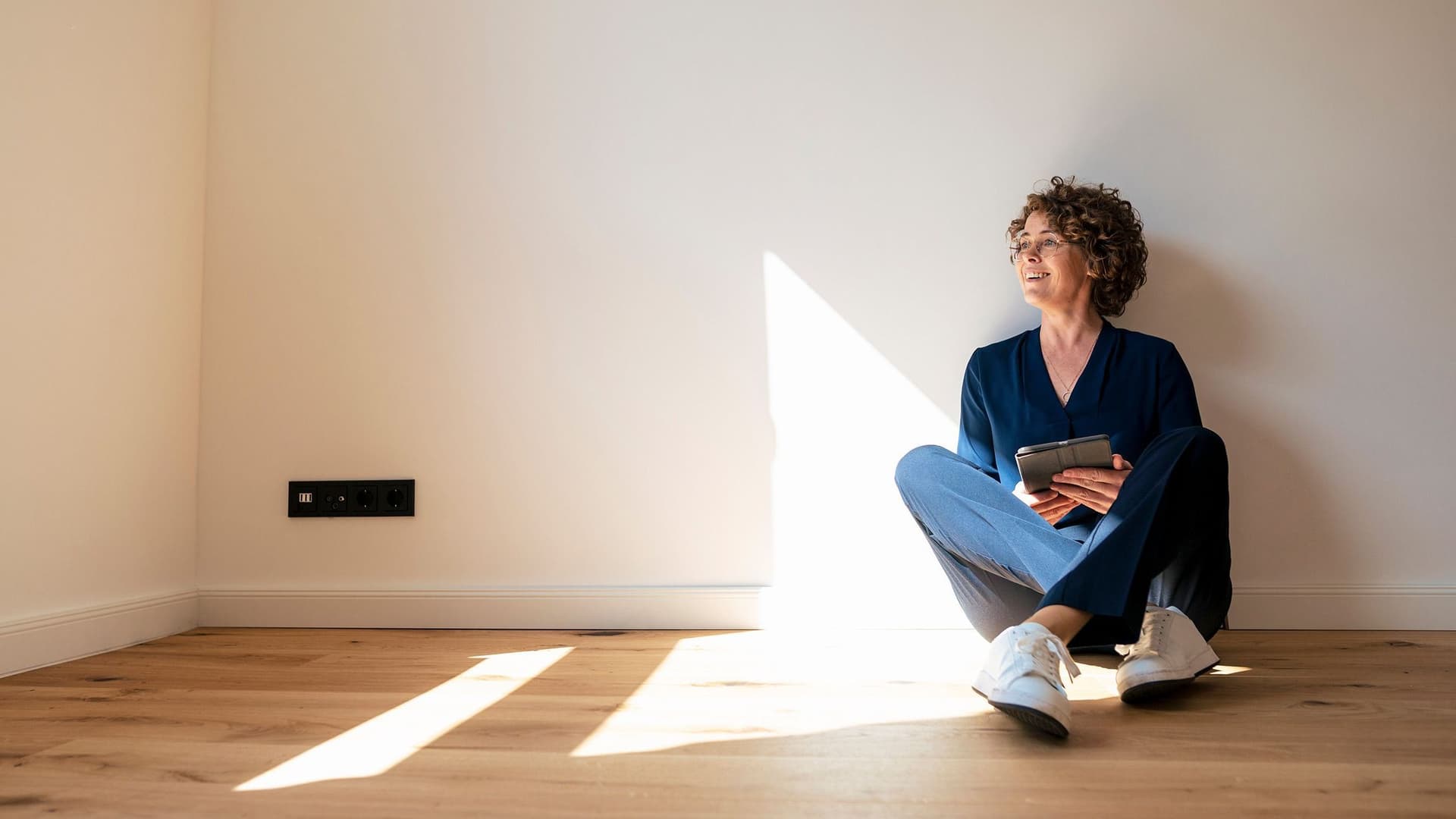 Eine junge Frau in Alltagskleidung sitzt auf dem Boden einer leeren Wohnung. Sie lächelt und schaut in Richtung Fenster.