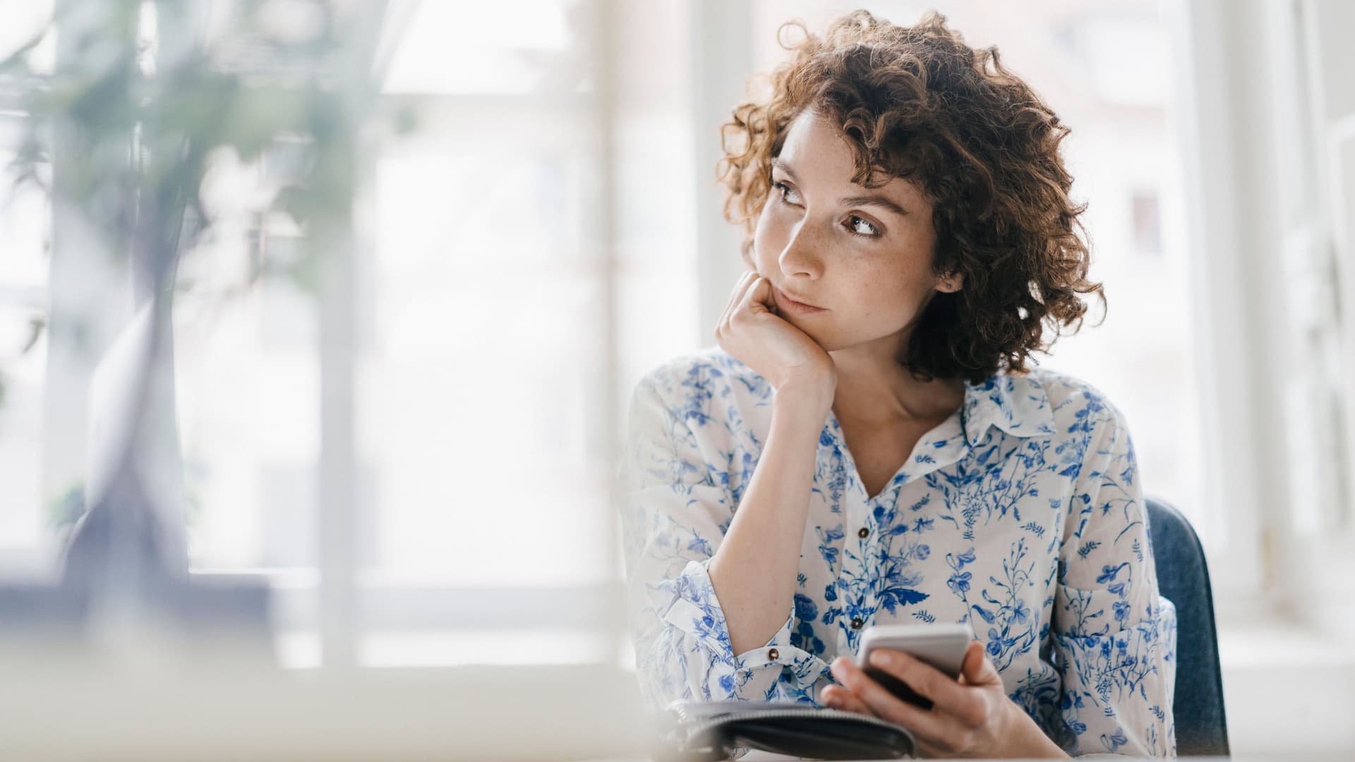 Junge Frau mit Smartphone in der Hand, hat ihren Kopf abgestützt und schaut verträumt zur Seite.