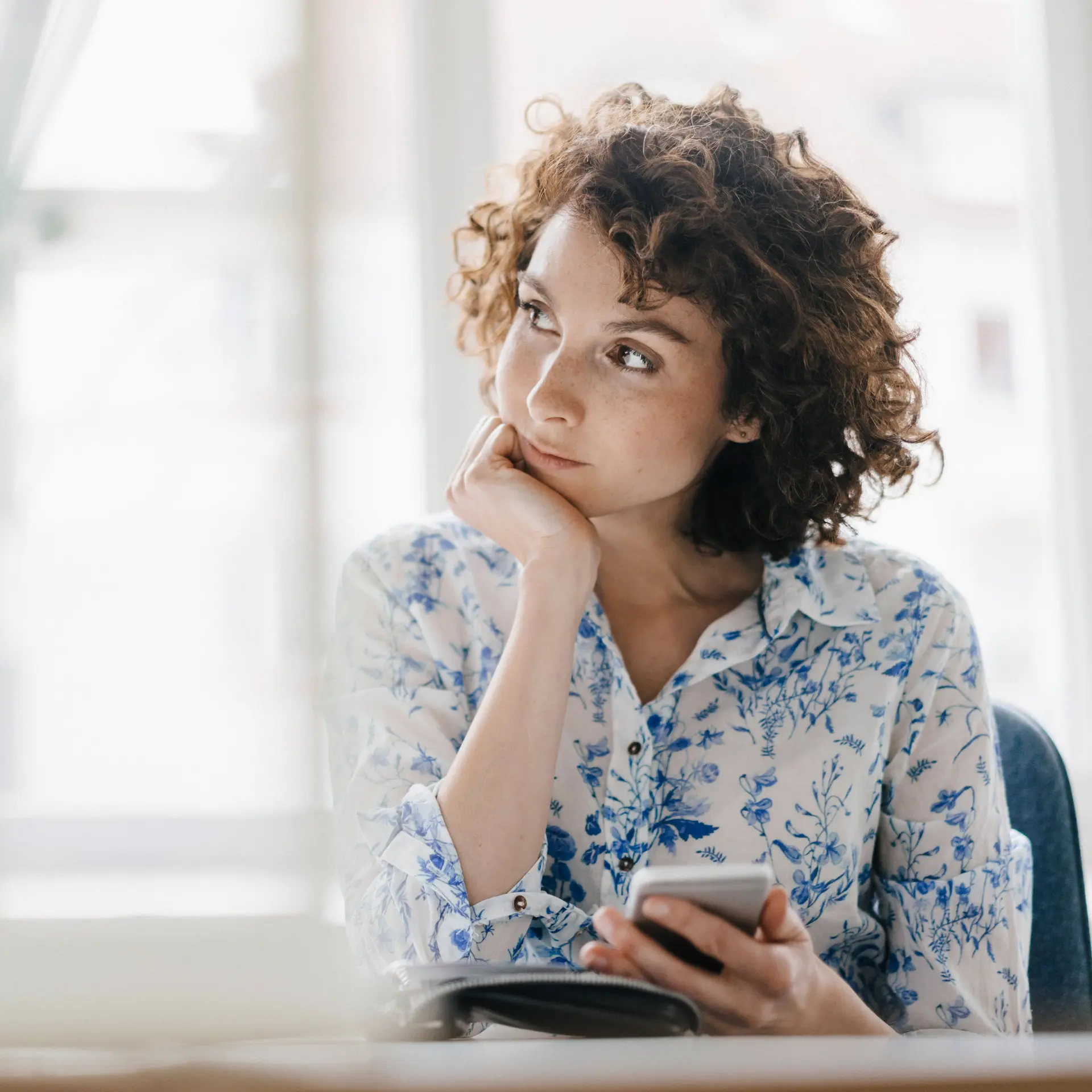 Junge Frau mit Smartphone in der Hand, hat ihren Kopf abgestützt und schaut verträumt zur Seite.
