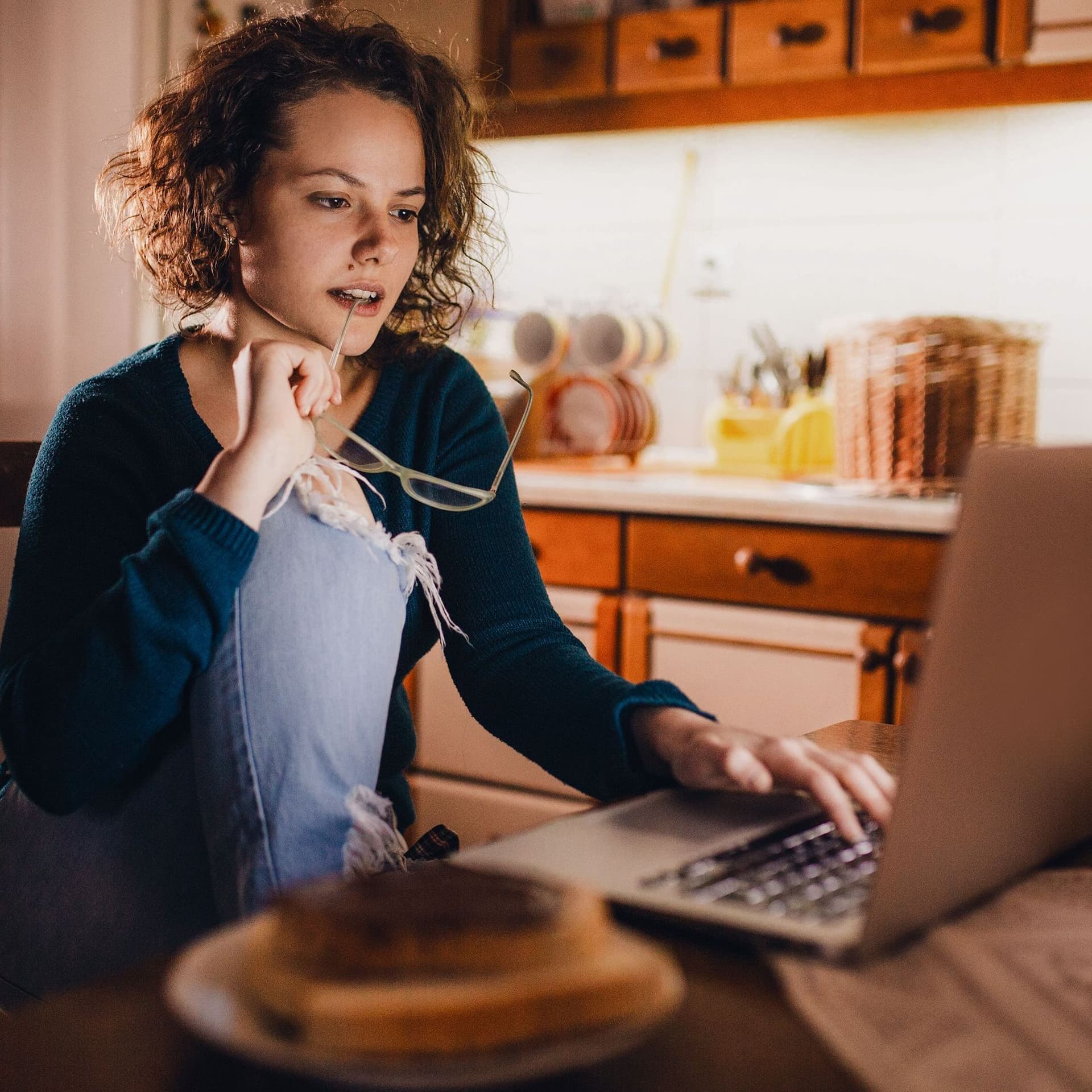 Eine Frau sitzt im Abendlicht an einem Tisch in ihrer Küche. Vor ihr steht ein Laptop. Sie hat ein Knie angewinkelt und beißt auf ihren Brillenbügel.