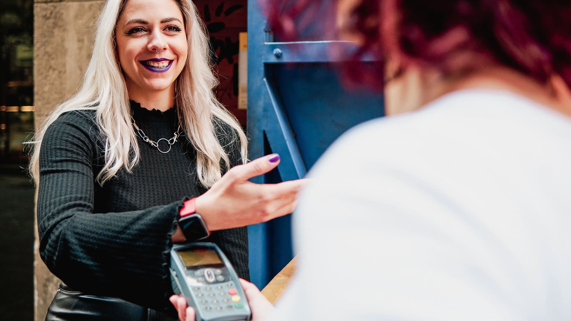 Blick über die Schulter einer Frau mit Kartenlesegerät. Frau mit schwarzem Lippenstift zahlt mit ihrer Smartwatch.