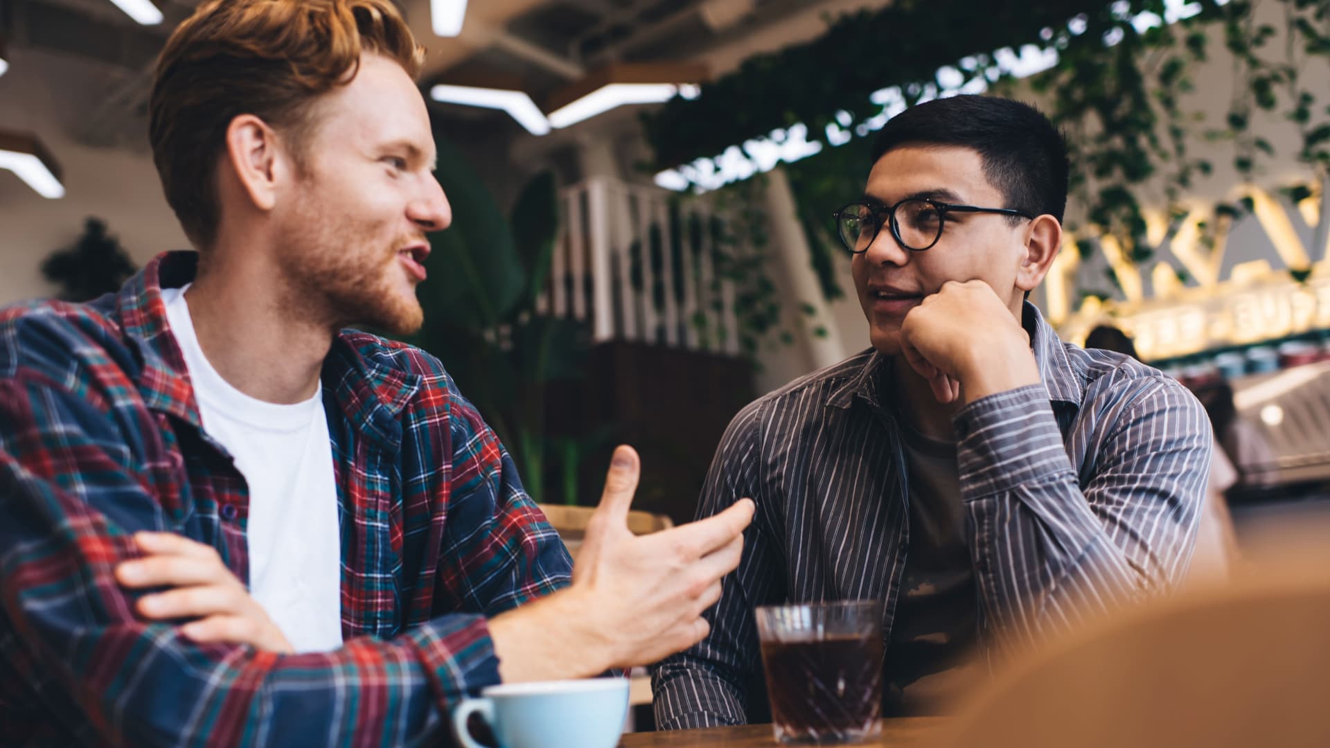 Junge Männer im Gespräch in einem Cafe. Auf einem Holztisch vor ihnen steht eine Tasse und ein Glas.