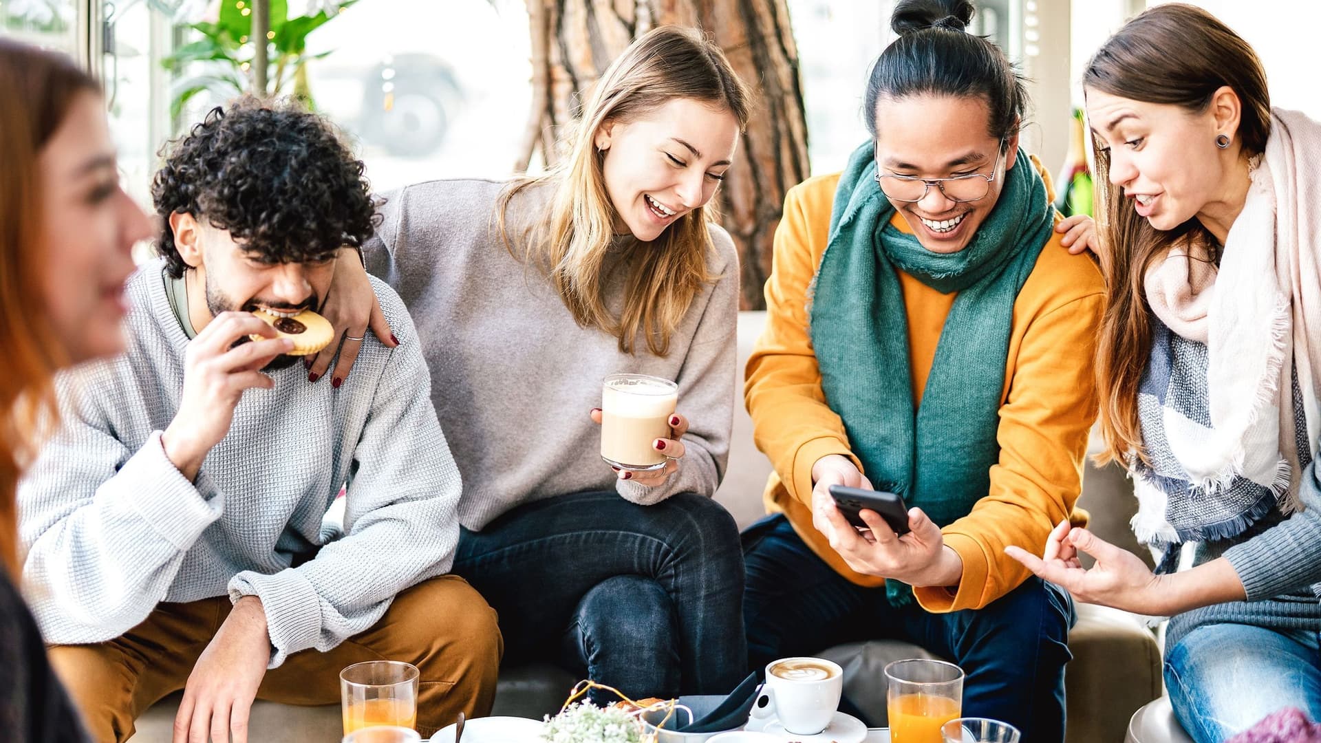 Freunde sitzen beim Essen und lachen. Einer hält ein Smartphone in der Hand.