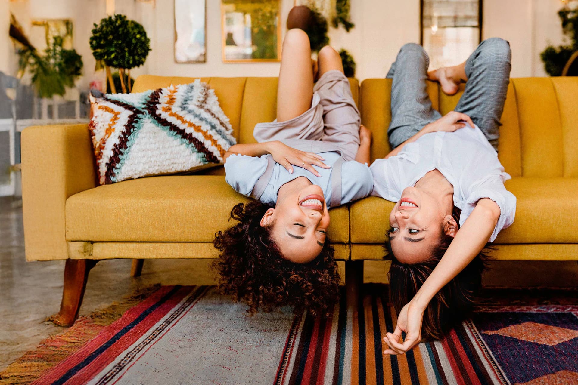 Zwei junge Frauen liegen rücklings und kopfüber auf einem gelben Sofa in einem Wohnzimmer. Sie lachen ausgelassen.