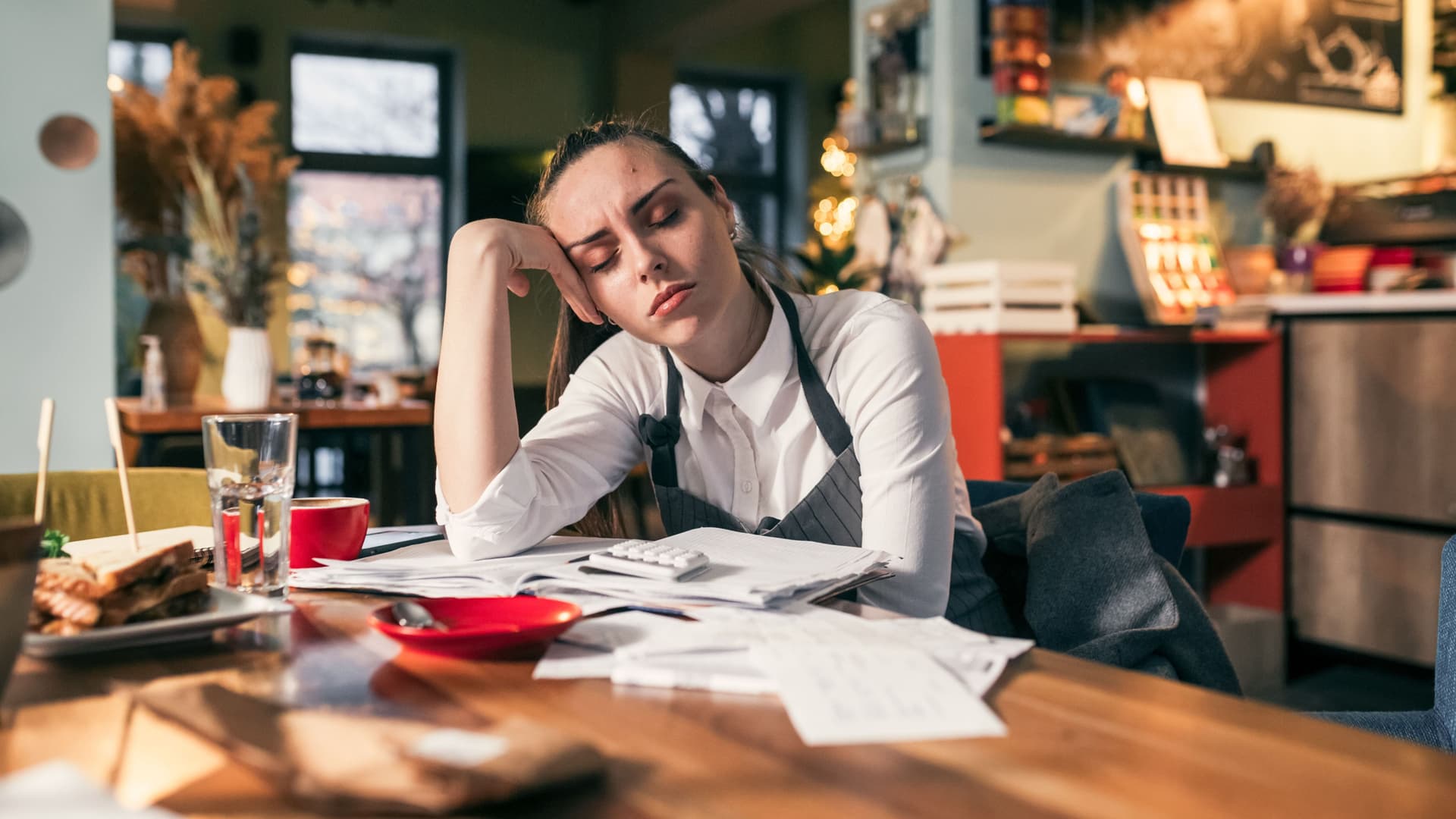 Eine traurig aussehende junge Frau aus der Gastronomie sitzt mit geschlossenen Augen über Rechnungen an einem Tisch in gemütlicher Atmosphäre.