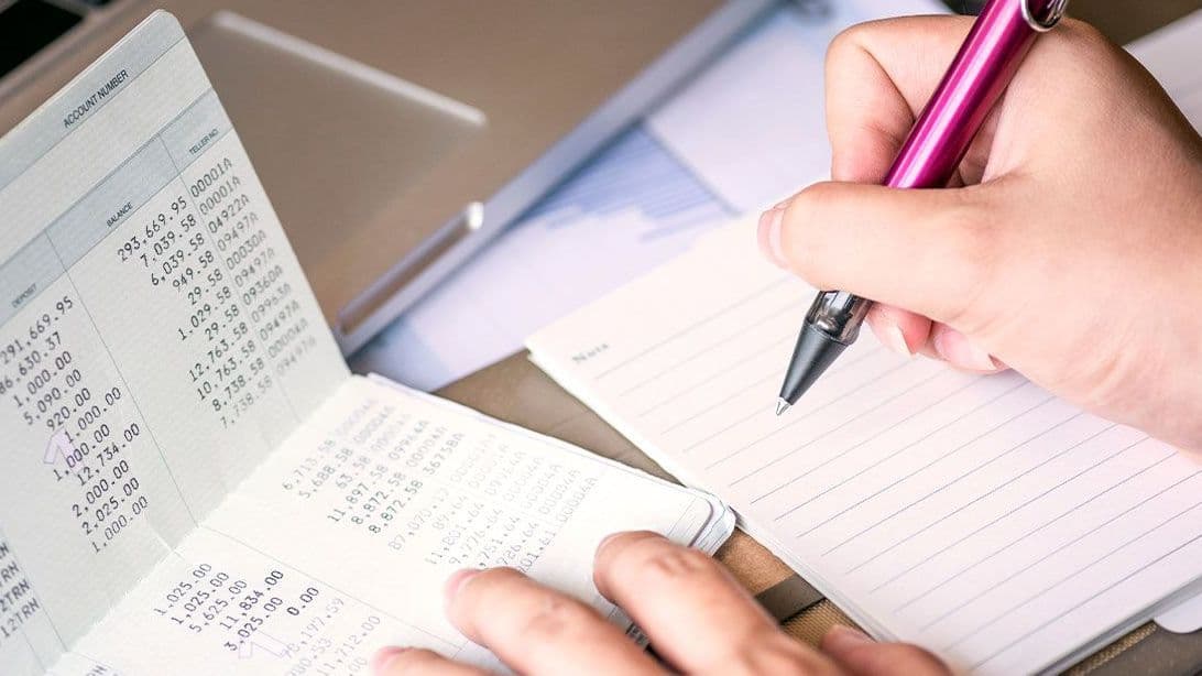 Nahaufnahme einer Hand, die einen magentafarbenen Kugelschreiber über einem Notizzettel hält und etwas aus einem Sparbuch notiert.