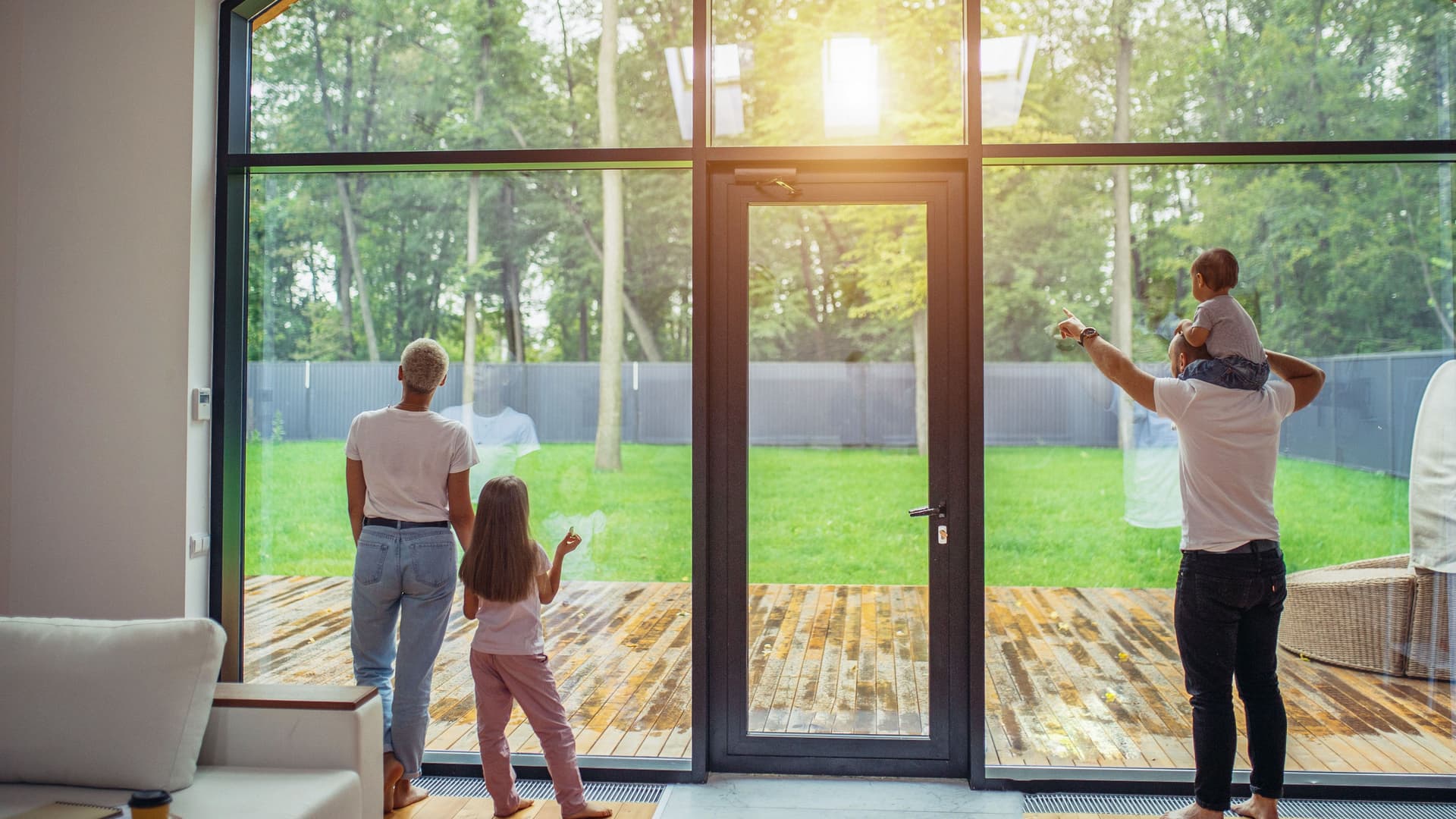 Eltern mit Kindern stehen vor einem großen Panoramafenster mit Tür und gucken in den Garten. Das Baby sitzt auf den Schultern des Vaters und die Tochter steht neben der Mutter.