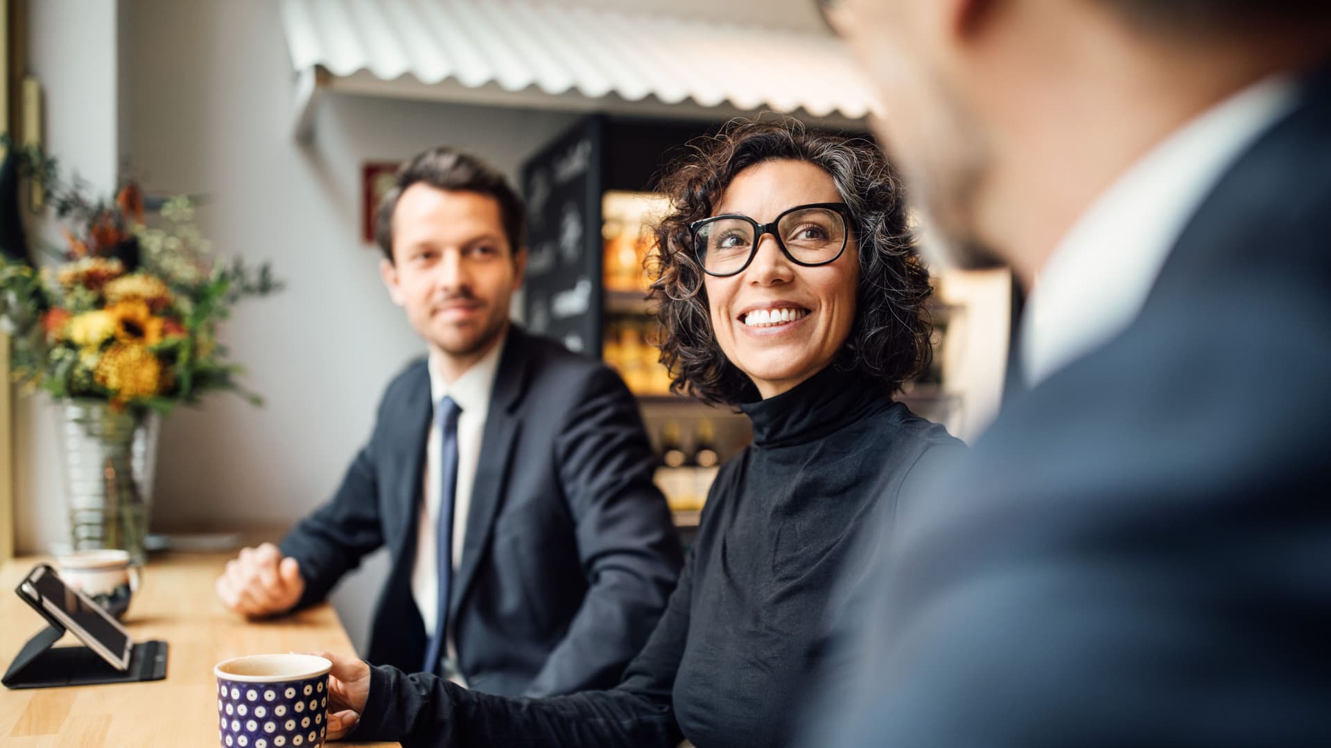 Ein Mann und eine Frau in Business-Kleidung sitzen in einem Cafe zusammen an einem langen Tisch. Sie lächeln und gucken einen anderen Mann an, den man nur zum Teil sieht.
