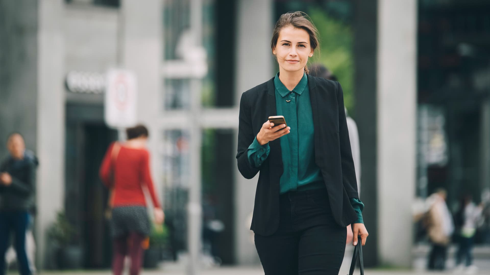 Junge Frau in Business-Kleidung mit Mobiltelefon in einer Hand und einer Tasche in der anderen, läuft auf einer Straße und blickt in die Kamera.