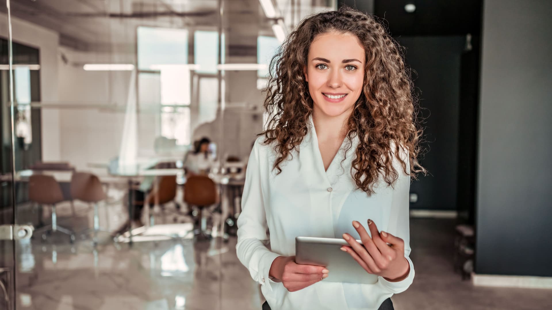 Eine junge Frau steht mit Tablet in der Hand in einem modernen Büro und blickt in die Kamera.