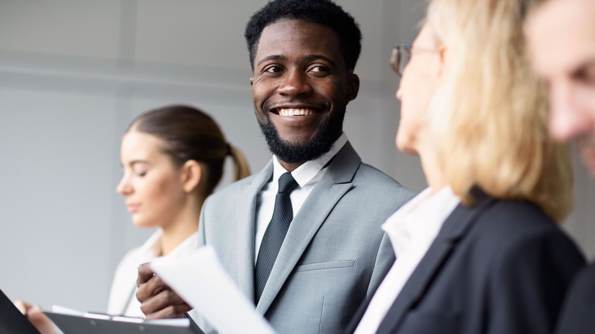 Interkulturelle Kompetenz Junger, lachender Mann in Anzug, der eine Kollegin ansieht. Eine andere Kollegin notiert sich etwas. Alle halten Mappen in der Hand.