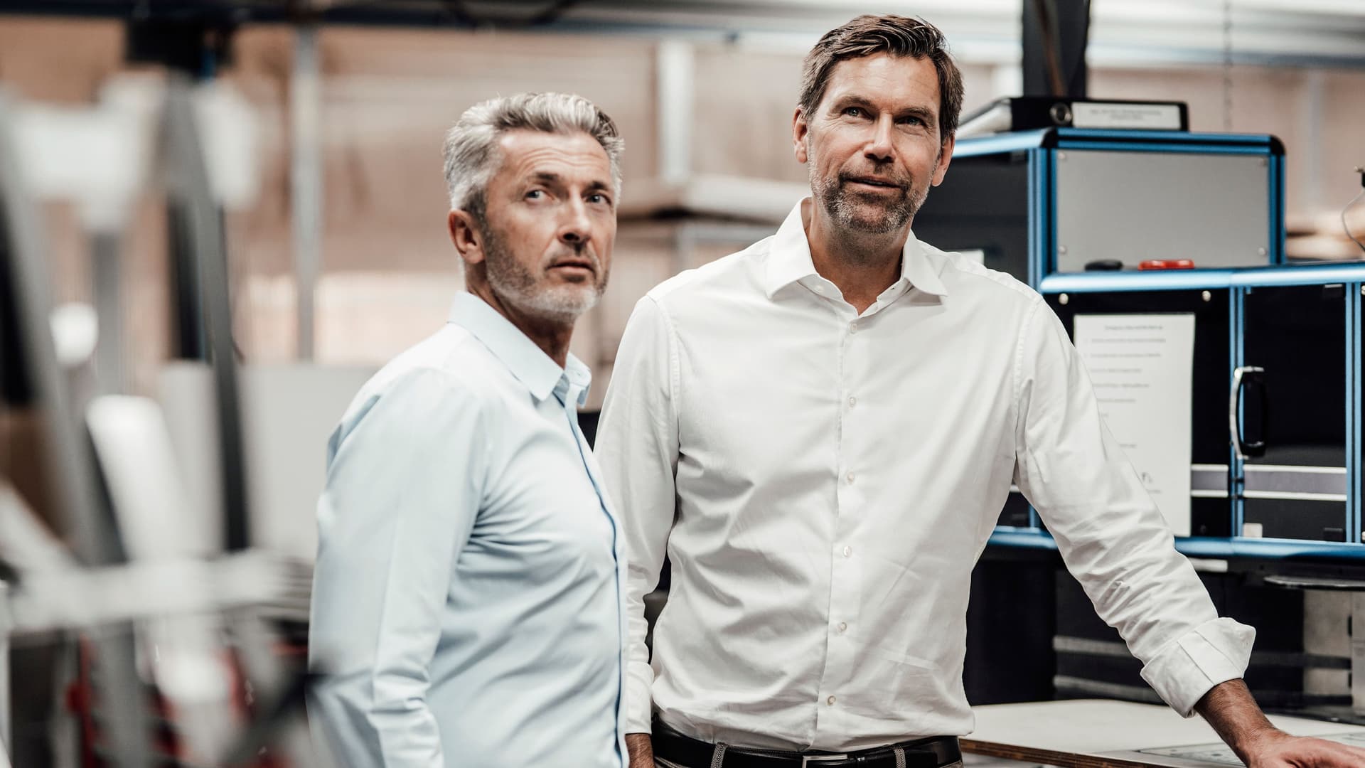 Zwei Männer mit Händen in den Taschen stehen in einer Produktionshalle. Sie tragen Hemden und gucken auf etwas.