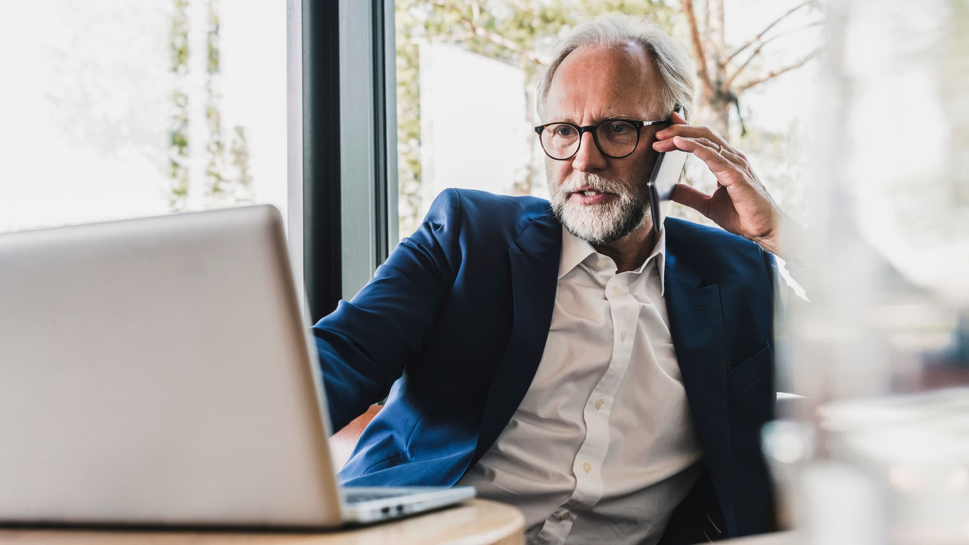 Ältere Mann in Anzug mit Handy am Ohr sitzt mit einem Laptop an einem Tisch in einem Café.