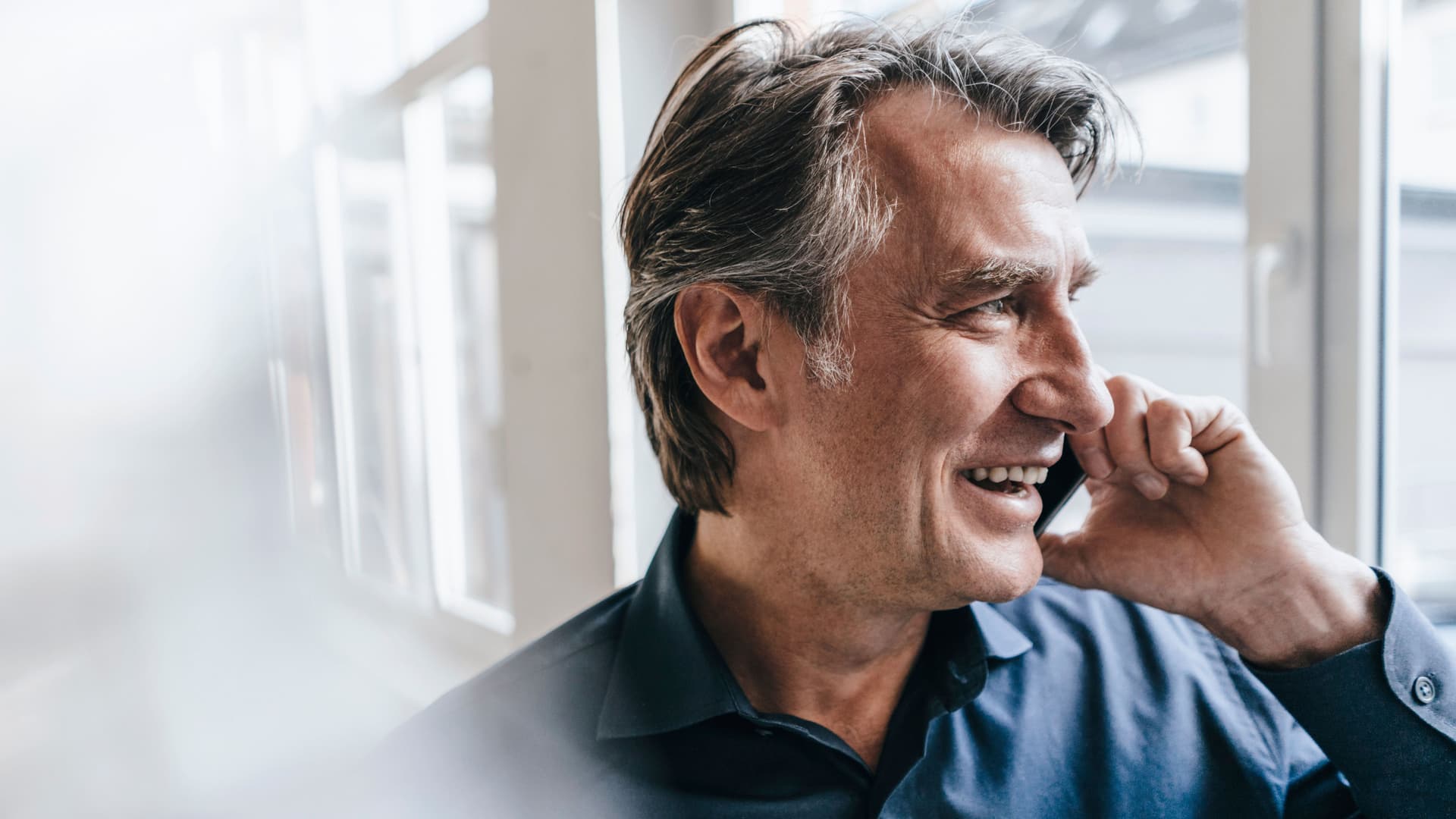 Lächelnder Mann mit Handy am Ohr. Er trägt ein Hemd und guckt aus einem Fenster.