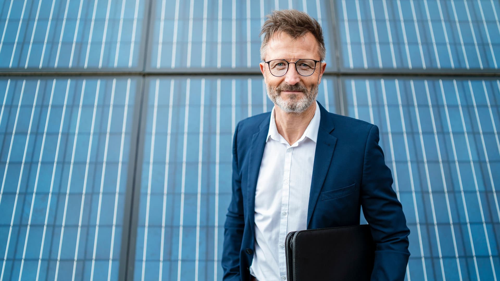 Porträt eines lächelnden Mannes, der einen Anzugjacke trägt und einen Ordner unter dem Arm hält. Er steht vor Solarzellen.