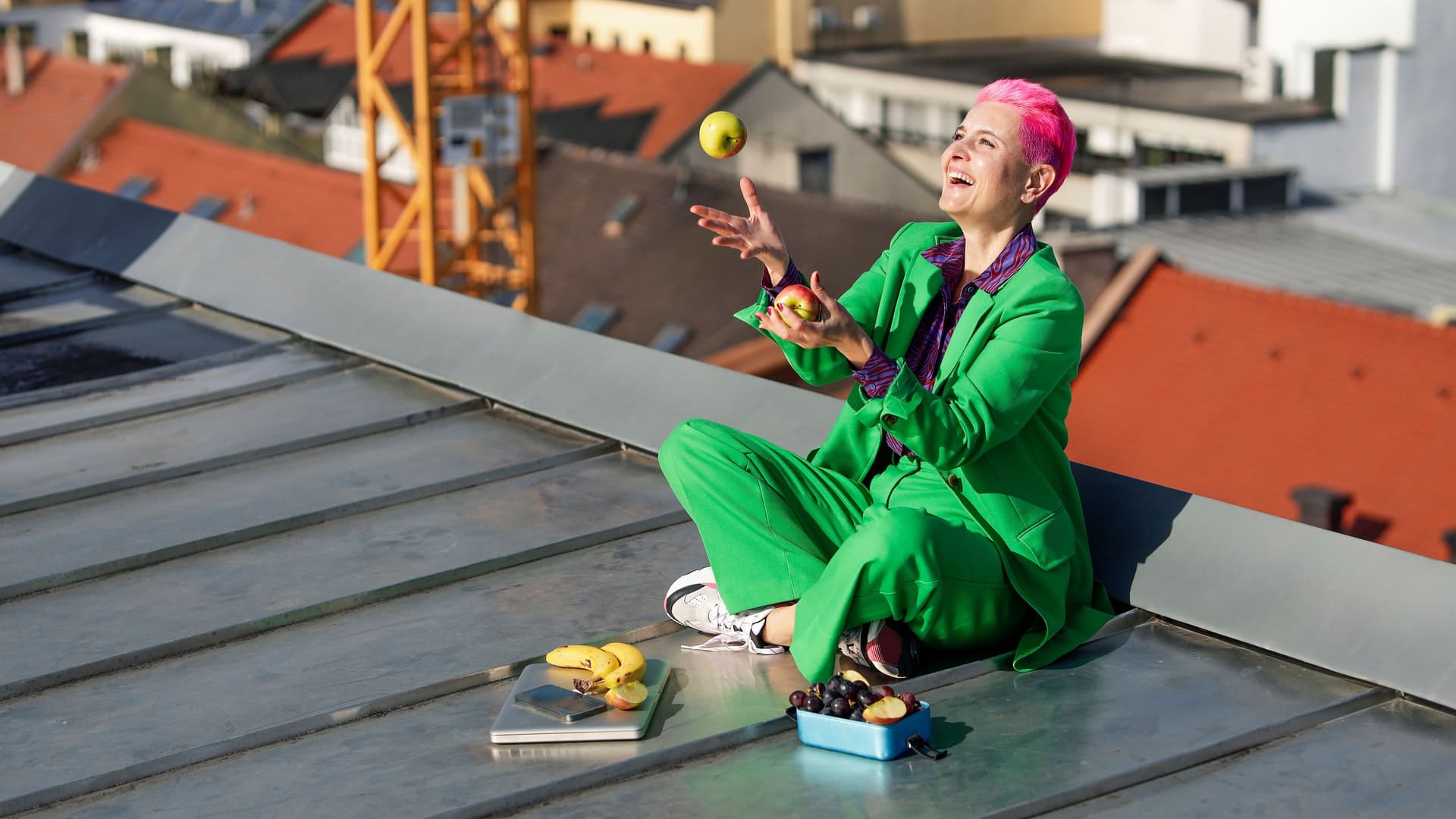 Lachende Frau sitzt auf einem Dach und jongliert mit zwei Äpfeln. Vor ihr liegt ein zugeklappter Laptop und mehr Obst in einer Dose.