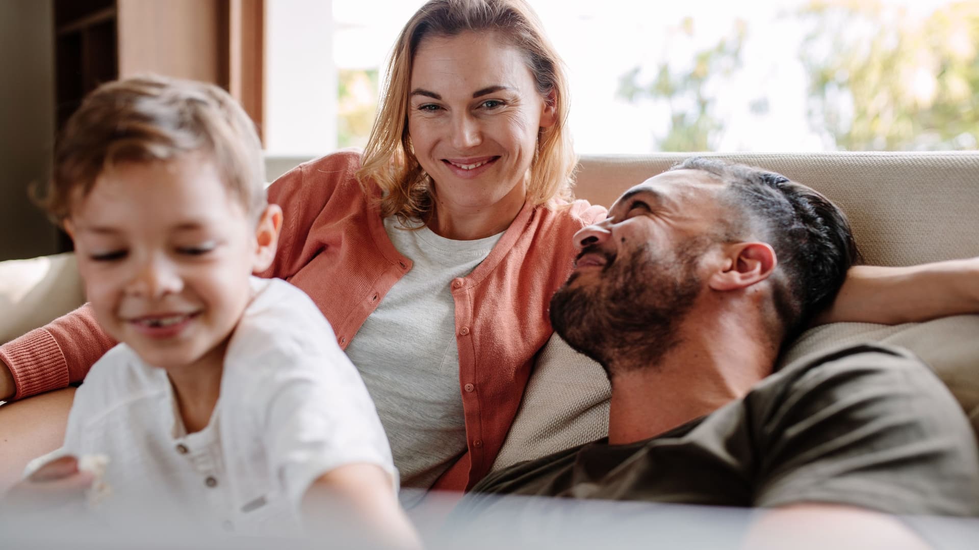 Eine junge Frau entspannt mit ihrem Mann und Sohn lächelnd auf einer Couch.