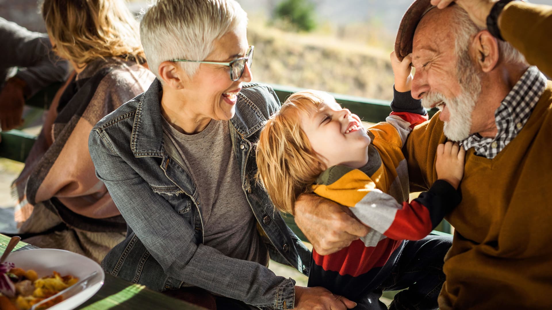 Lachende Großeltern albern mit Enkelkind während eines Restaurantbesuchs. Sie sitzen im Freien auf einem Balkon bei Sonnenschein.