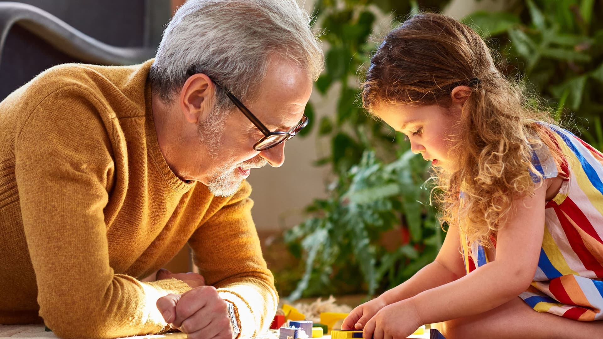Enkelin spielt mit Holzklötzen auf dem Boden und Opa schaut zu.
