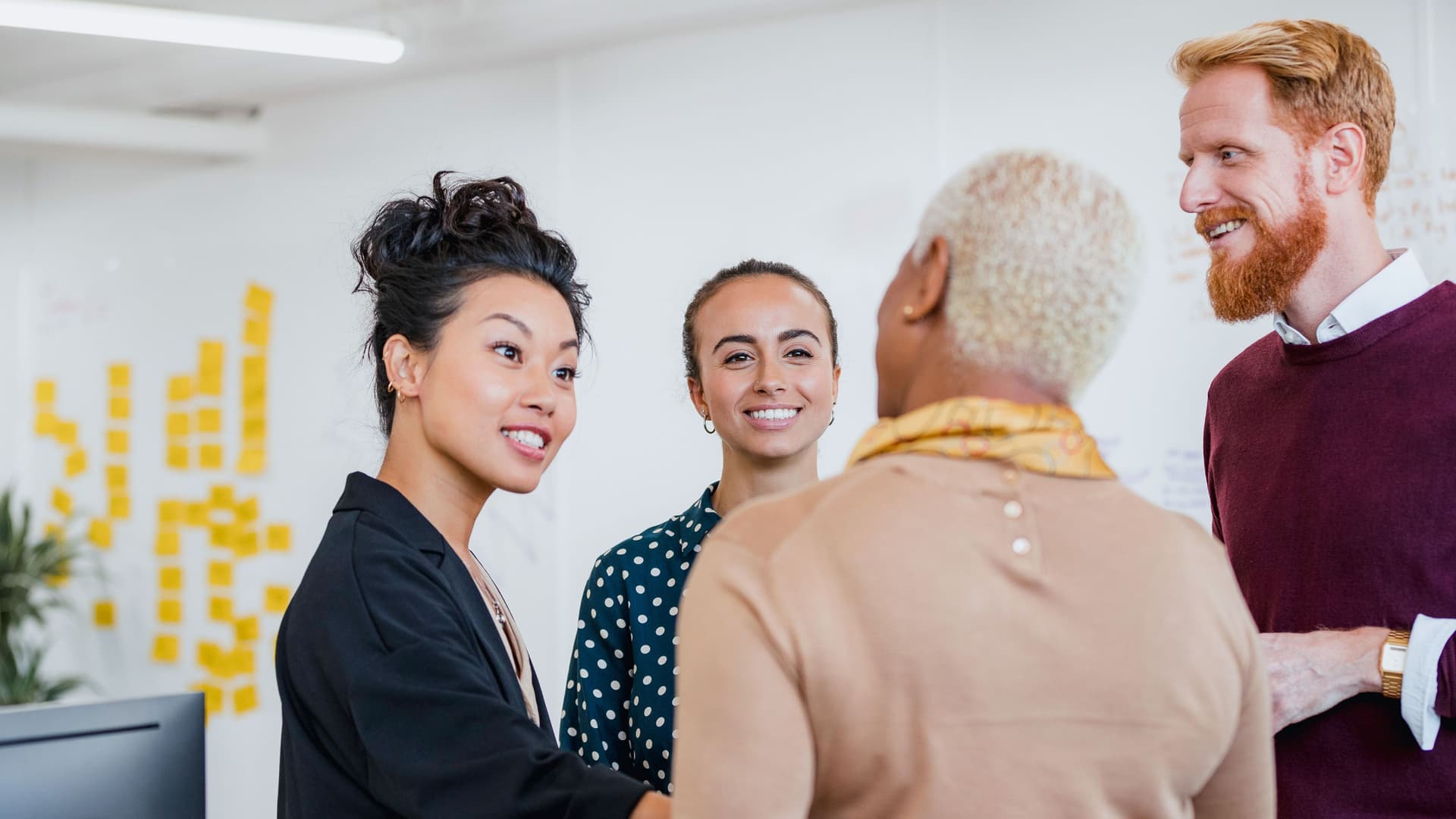Vier Personen stehen in einem Büro in einem Kreis und unterhalten sich. Im Hintergrund sieht man gelbe Haftnotizen an einer Wand kleben.