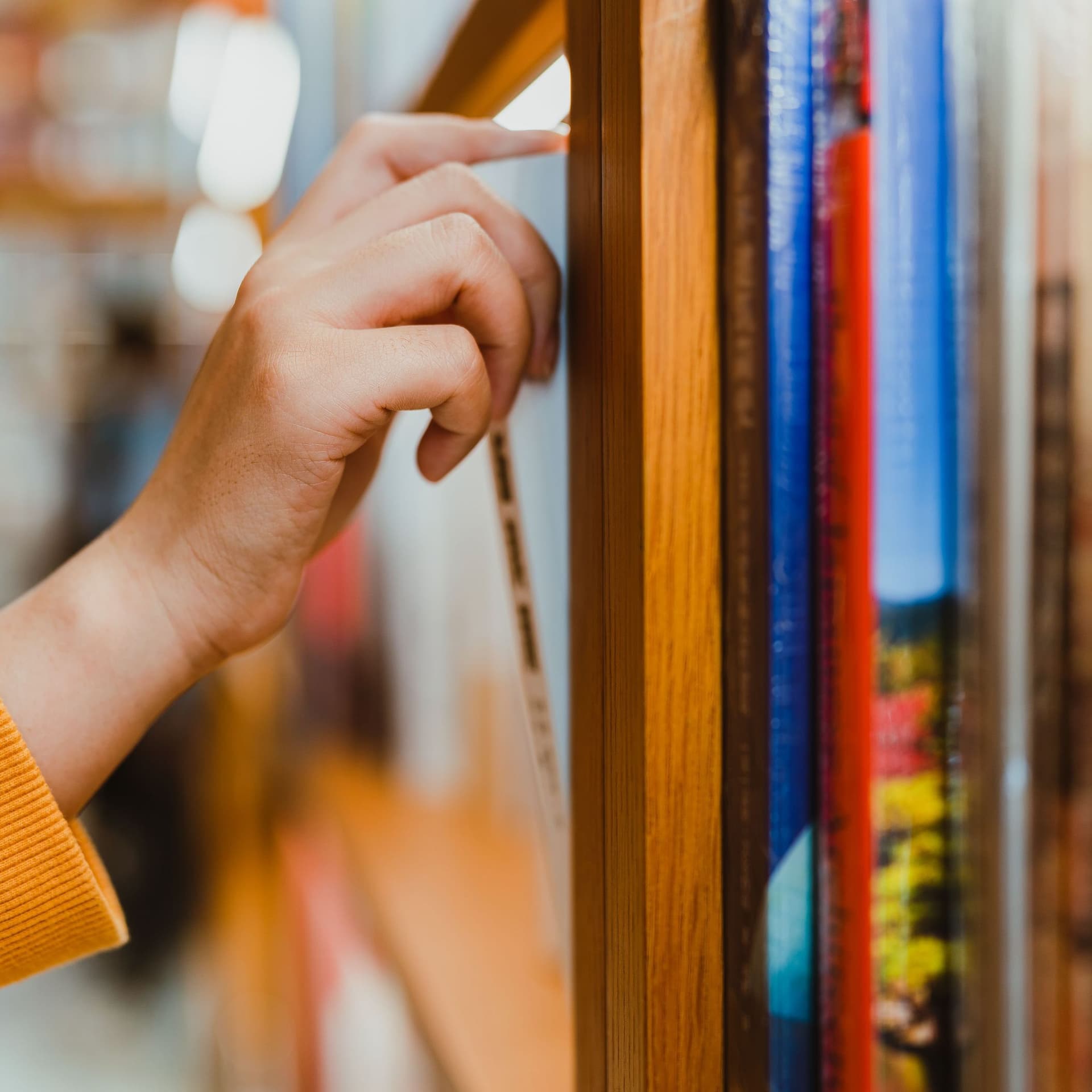 Seitliche Nahaufnahme von einem Arm mit orangefarbenen Pullover. Die Hand zieht ein Buch aus einem Bücherregal.