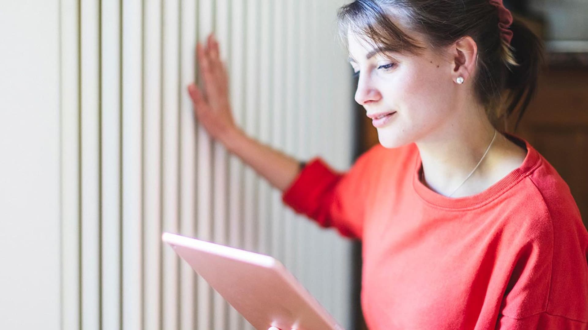 Eine junge Frau mit einem Tablet in der Hand fühlt die Temperatur eines Heizkörpers zuhause.