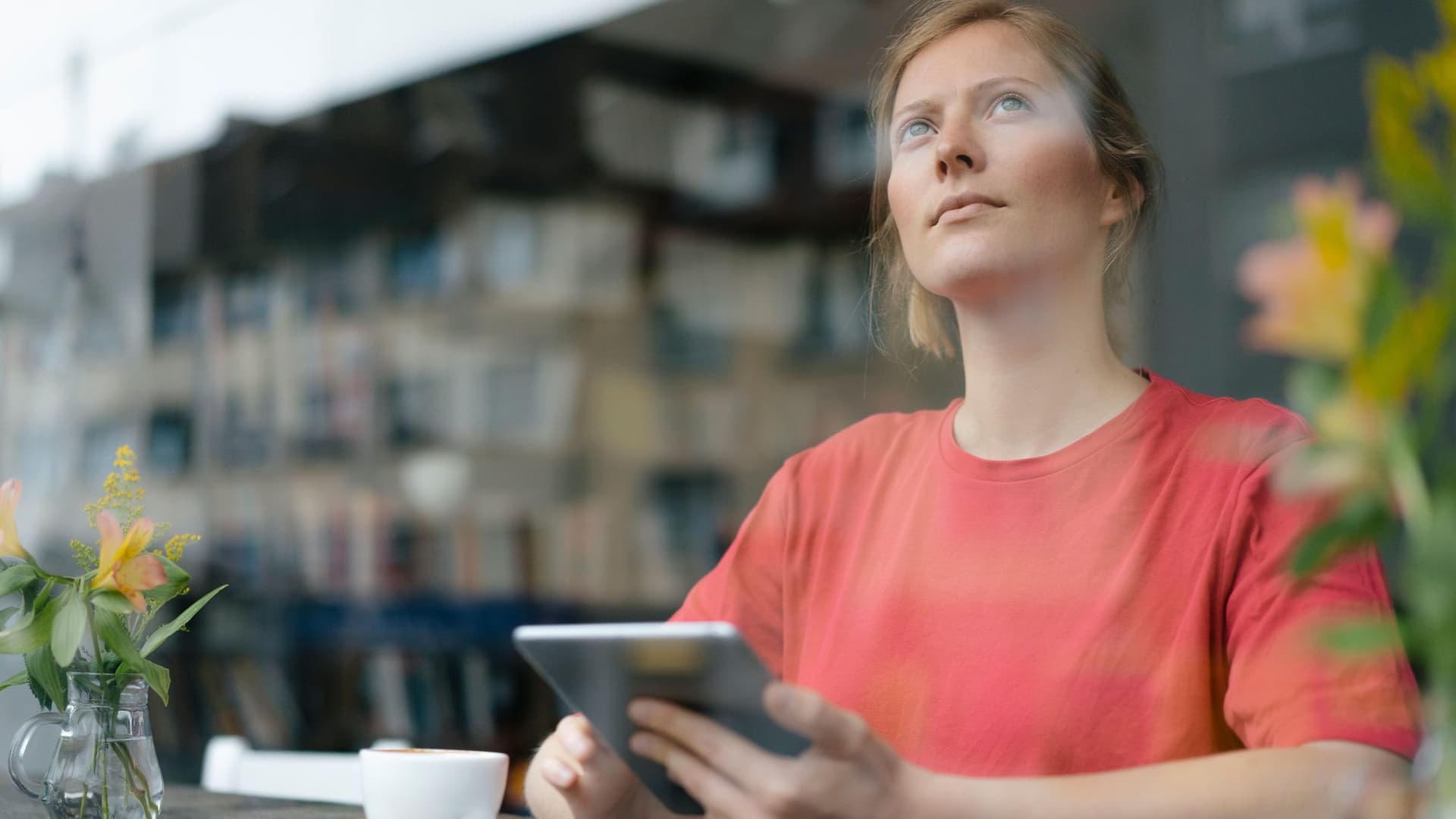 Eine junge Frau mit einem Tablet in der Hand sitzt in einem Café und schaut nach oben auf die Wohnhäuser gegenüber, die sich in der Fensterscheibe spiegeln.