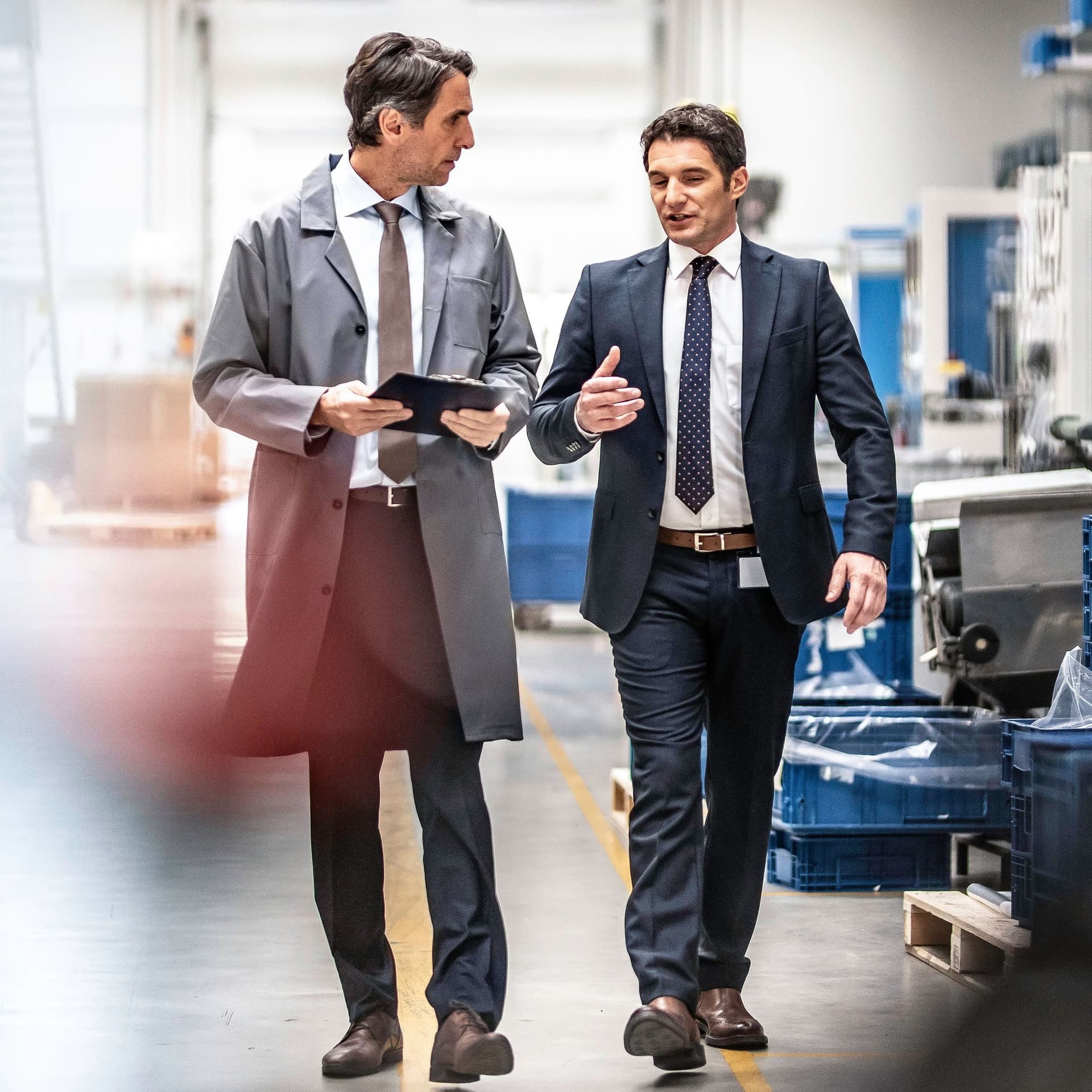 Ingenieur und Werkleiter laufen in Anzug durch eine Fabrik und reden miteinander.