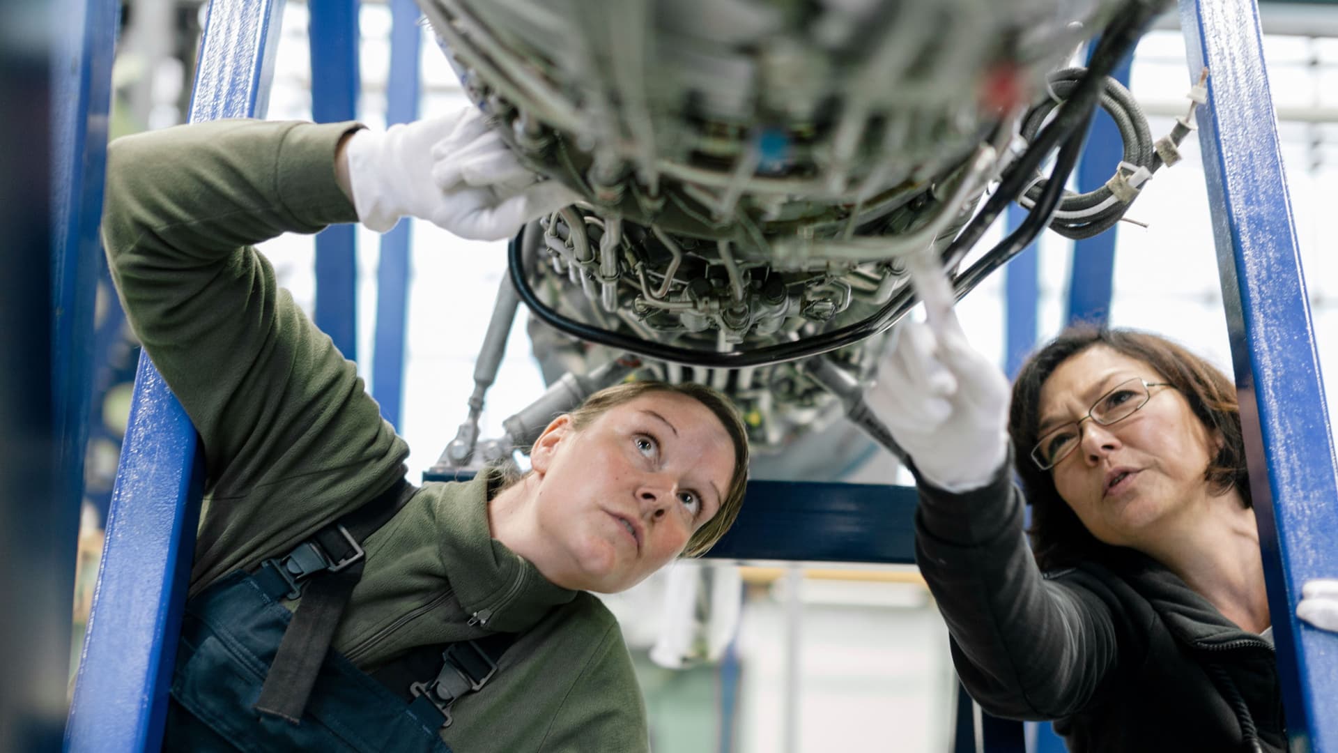 Zwei Mechanikerinnen untersuchen ein Flugzeugtriebwerk. Sie schauen von unten auf eine Vielzahl von Kabeln.