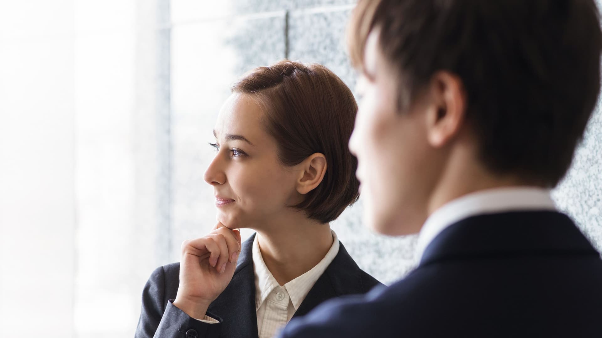 Entsendung Ausland Junge Frau in Business-Kleidung hat ihre Hand am Kinn und guckt zur Seite. Vor ihr steht ein Mann in Anzug mit dem Rücken zur Kamera.