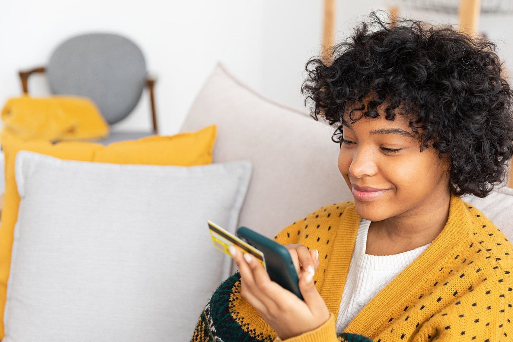 Eine junge Frau sitzt auf einem Sofa und hält neben einer goldenen Kreditkarte, ein Smartphone in den Händen. Sie schaut aufs Display.