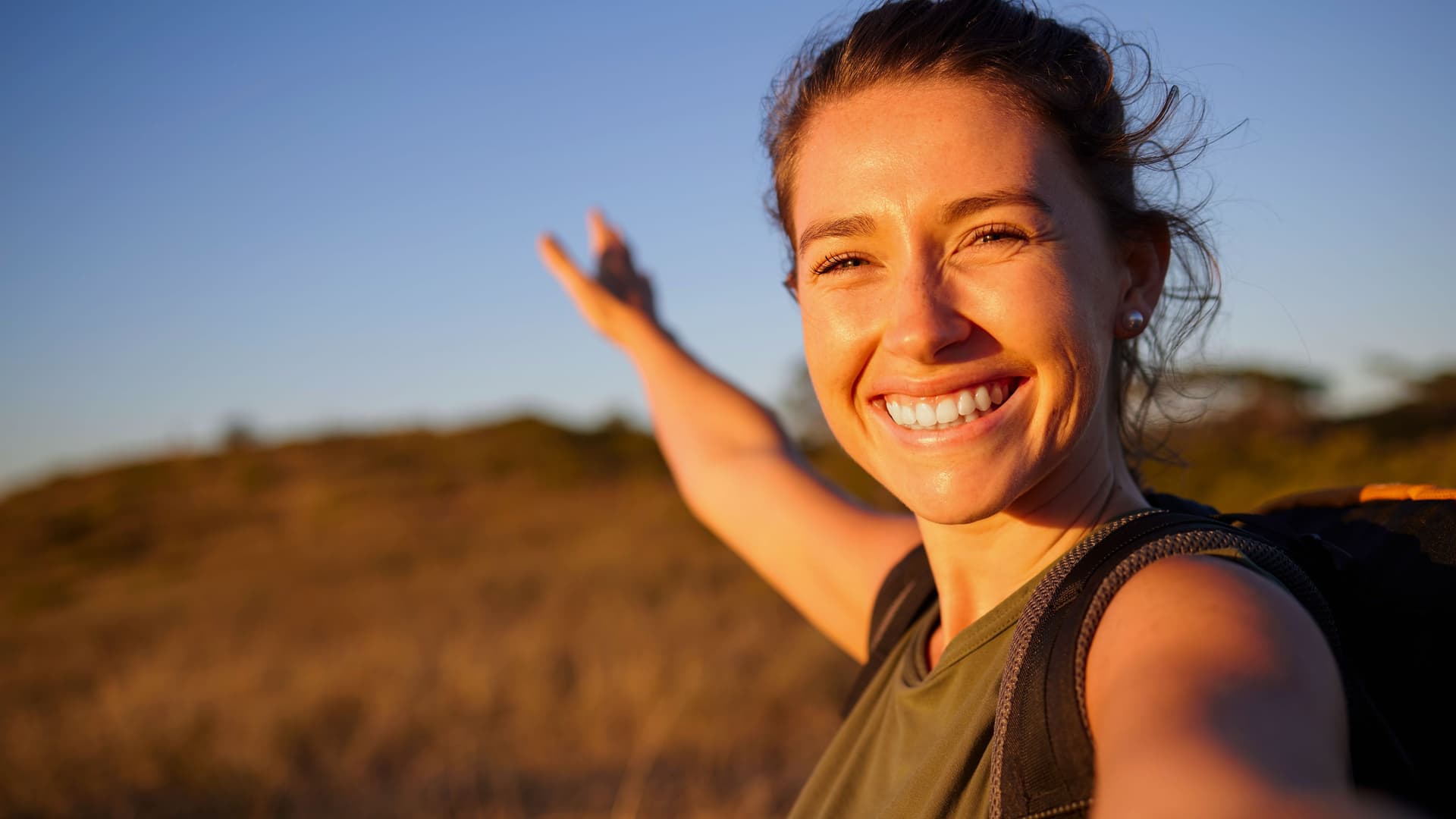 Lächelnde Frau mit Rucksack in tiefstehendem Sonnenlicht streckt einen Arm aus.