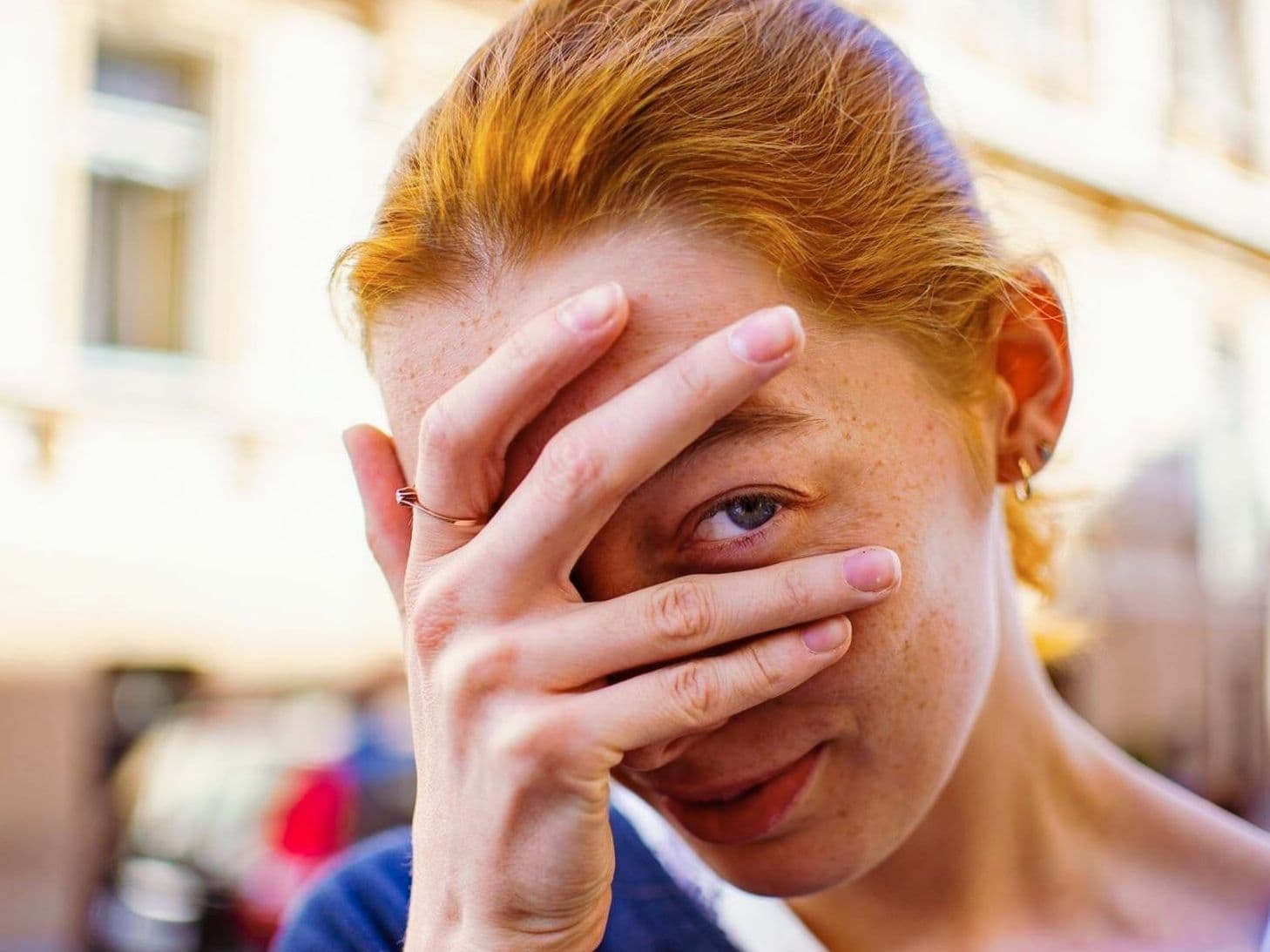 Straßenportrait einer jungen Frau, die ihr Gesicht mit ihrer Hand bedeckt. Sie schaut mit ihrem linken Auge zwischen Ring- und Mittelfinger hindurch.