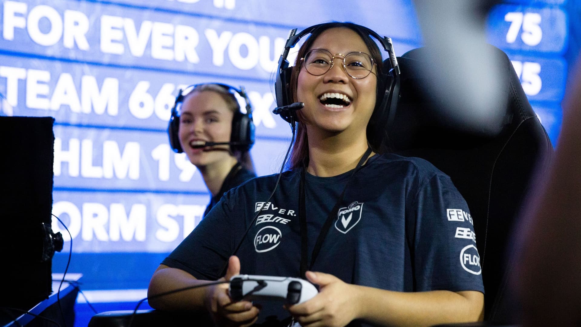 Eine junge Frau hält einen Controller in der Hand. Sie lacht. Im hintergrund ist eine beleuchtete Anzeigetafel.