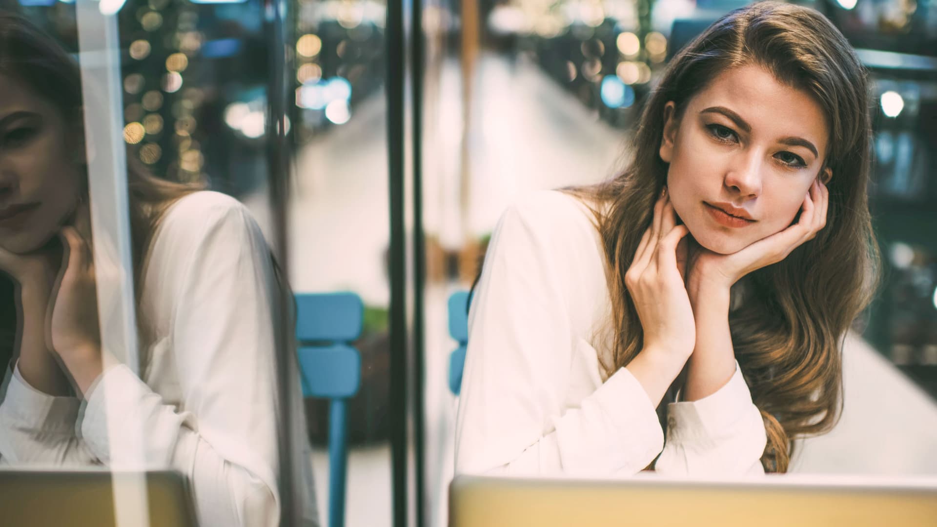 Porträt einer jungen Frau mit Laptop, die in die Kamera schaut. Links spiegelt sie sich in einer Glasscheibe.