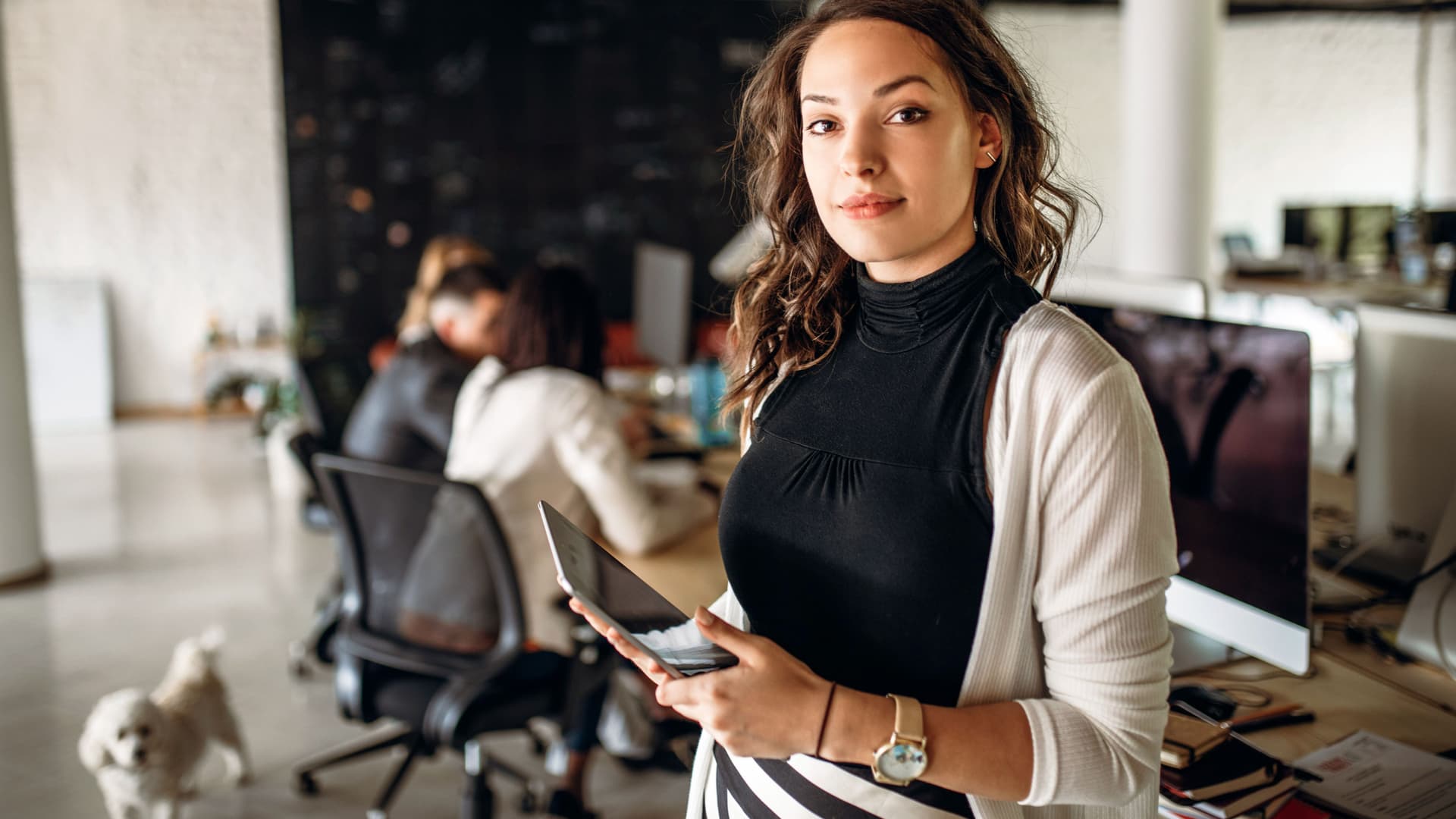 Eine junge Frau steht in einem modernen Büro uns schaut in die Kamera. Im Hintergrund ist verschwommen ein Team und ein Hund zu sehen.