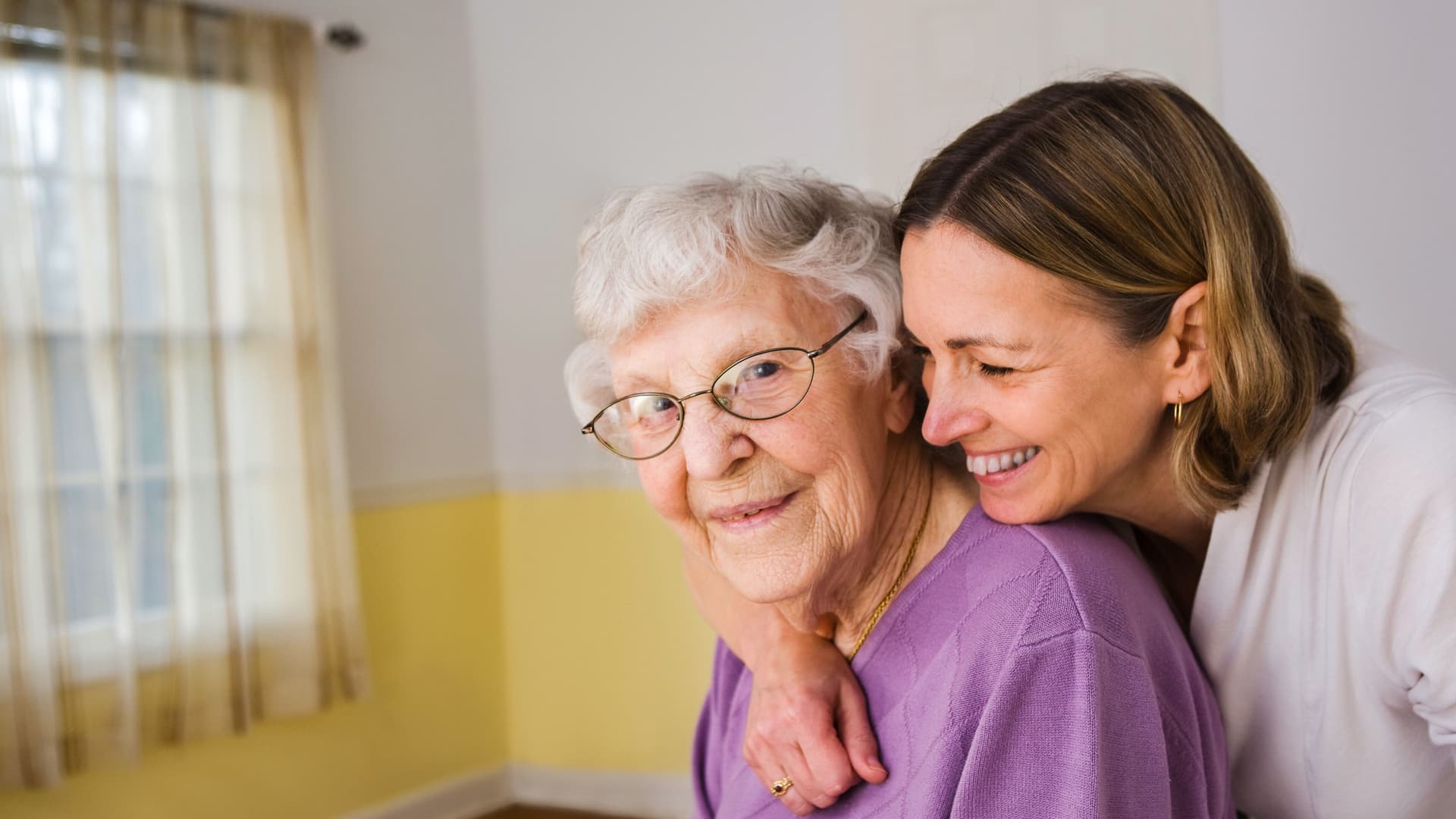 Eine ältere Frau mit grauen Haaren und Brille wird liebevoll von einer jüngeren Frau umarmt; beide lächeln in einem hellen, freundlich eingerichteten Zimmer.