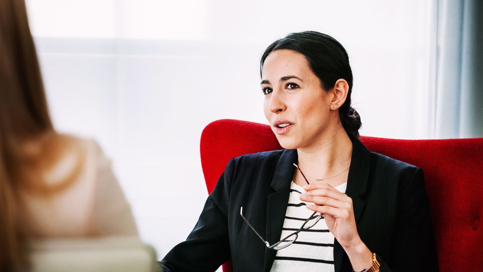 Frau mit Business-Kleidung und Brille in der Hand sitzt im Gespräch mit einer anderen Frau von der man nur einen Teilausschnitt von hinten sieht in einem Hotelzimmer.