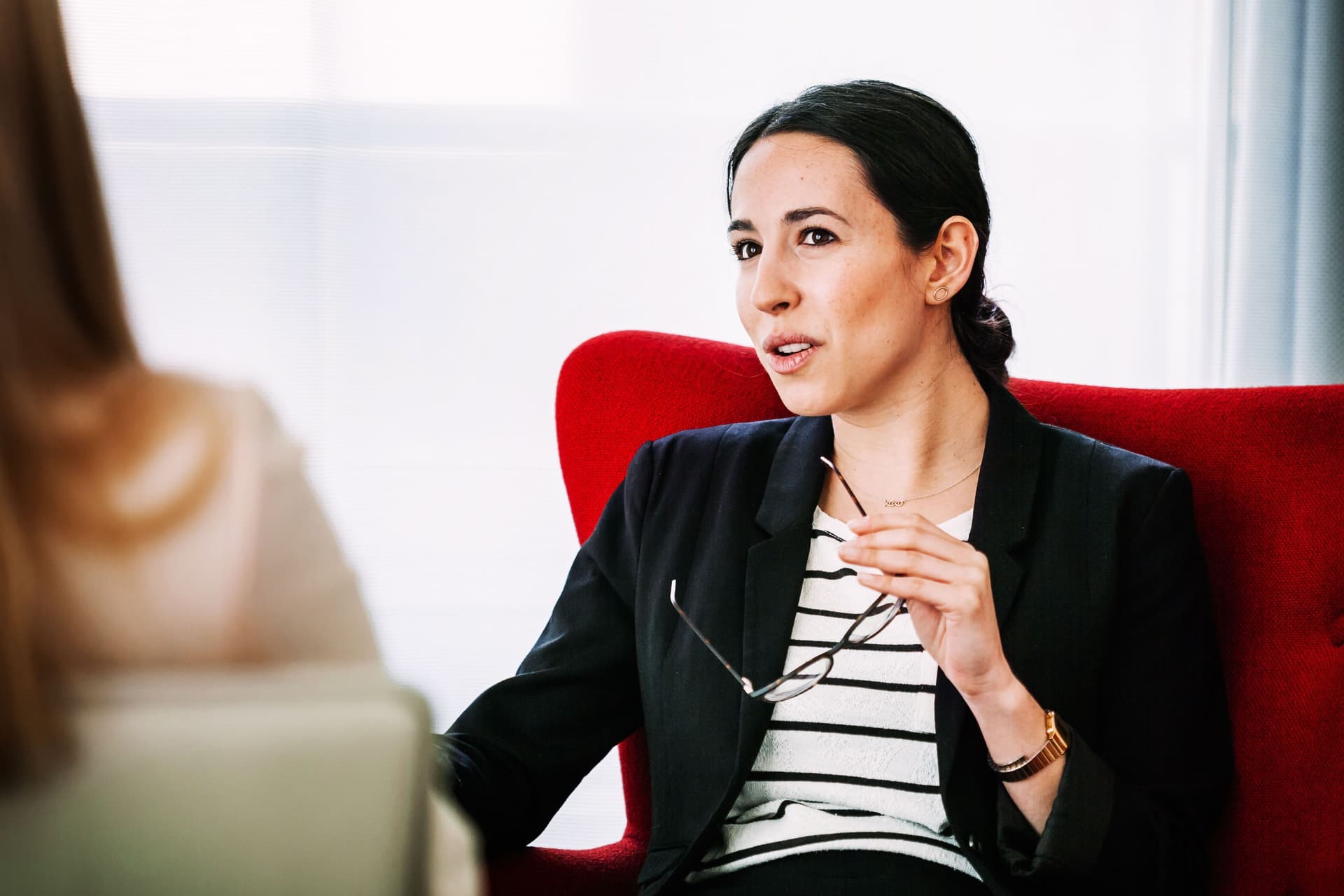Frau mit Business-Kleidung und Brille in der Hand sitzt im Gespräch mit einer anderen Frau von der man nur einen Teilausschnitt von hinten sieht in einem Hotelzimmer.