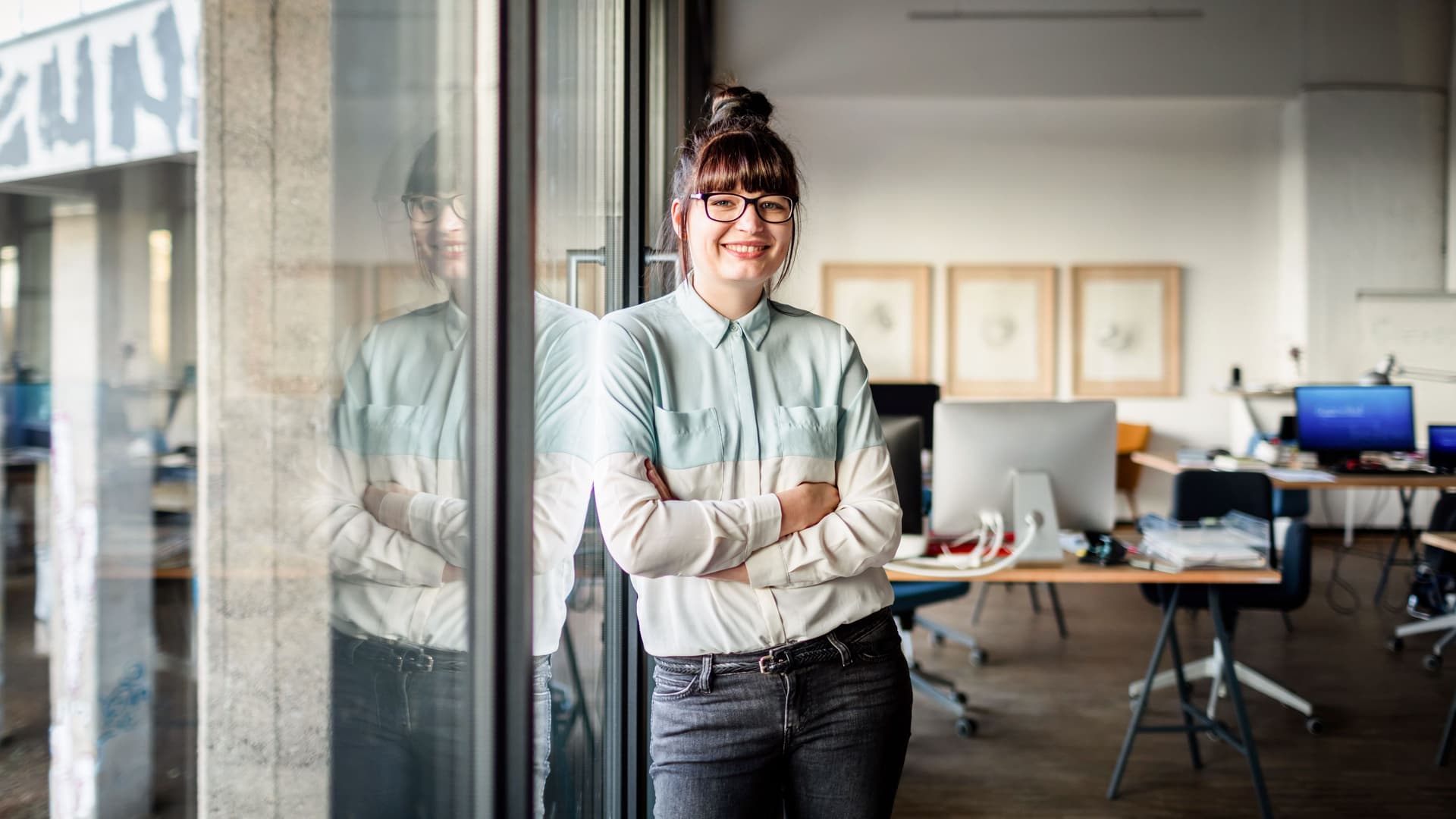 Junge Frau mit Brille und Dutt, die mit verschränkten Armen an einer Glasscheibe in einem modernen Büro lehnt und in die Kamera lächelt.