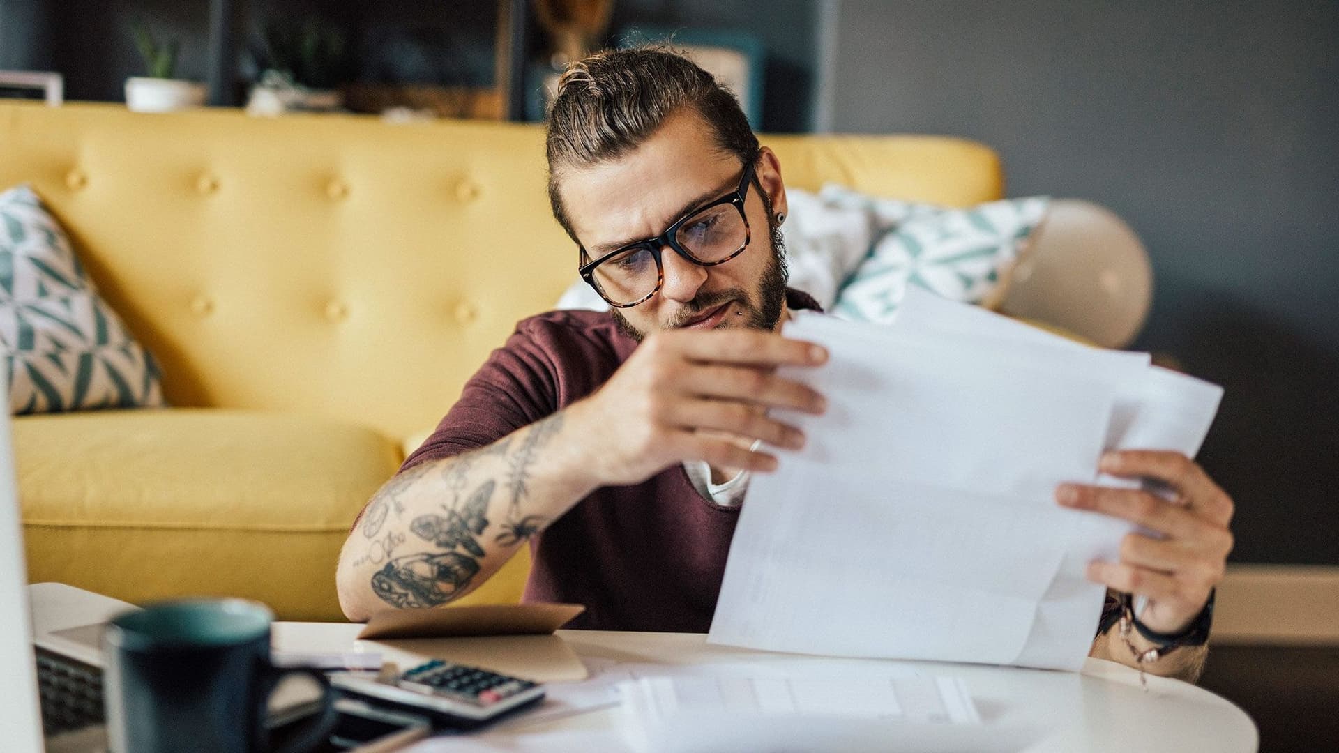 Ein junger Mann mit Brille und mittellange nach hinten gebundene Haare haelt konzentriert mehrere Dokumente in den Haenden.