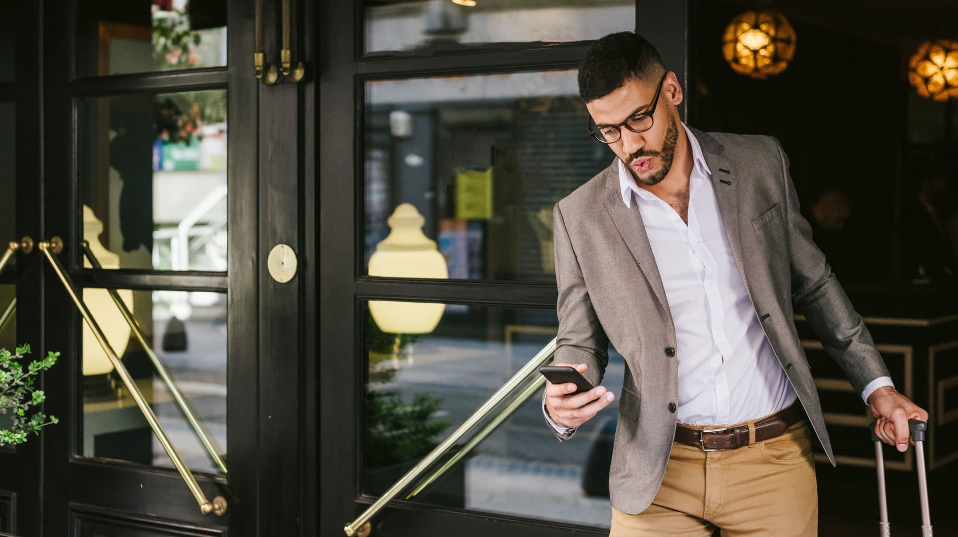 Junger Mann mit kurzen, dunklen Haaren, Brille und Bart zieht mit der einen Hand einen Rollkoffer und hält in der anderen ein Smartphone, während er ein Gebäude durch eine von mehreren verglasten Türen verlässt.