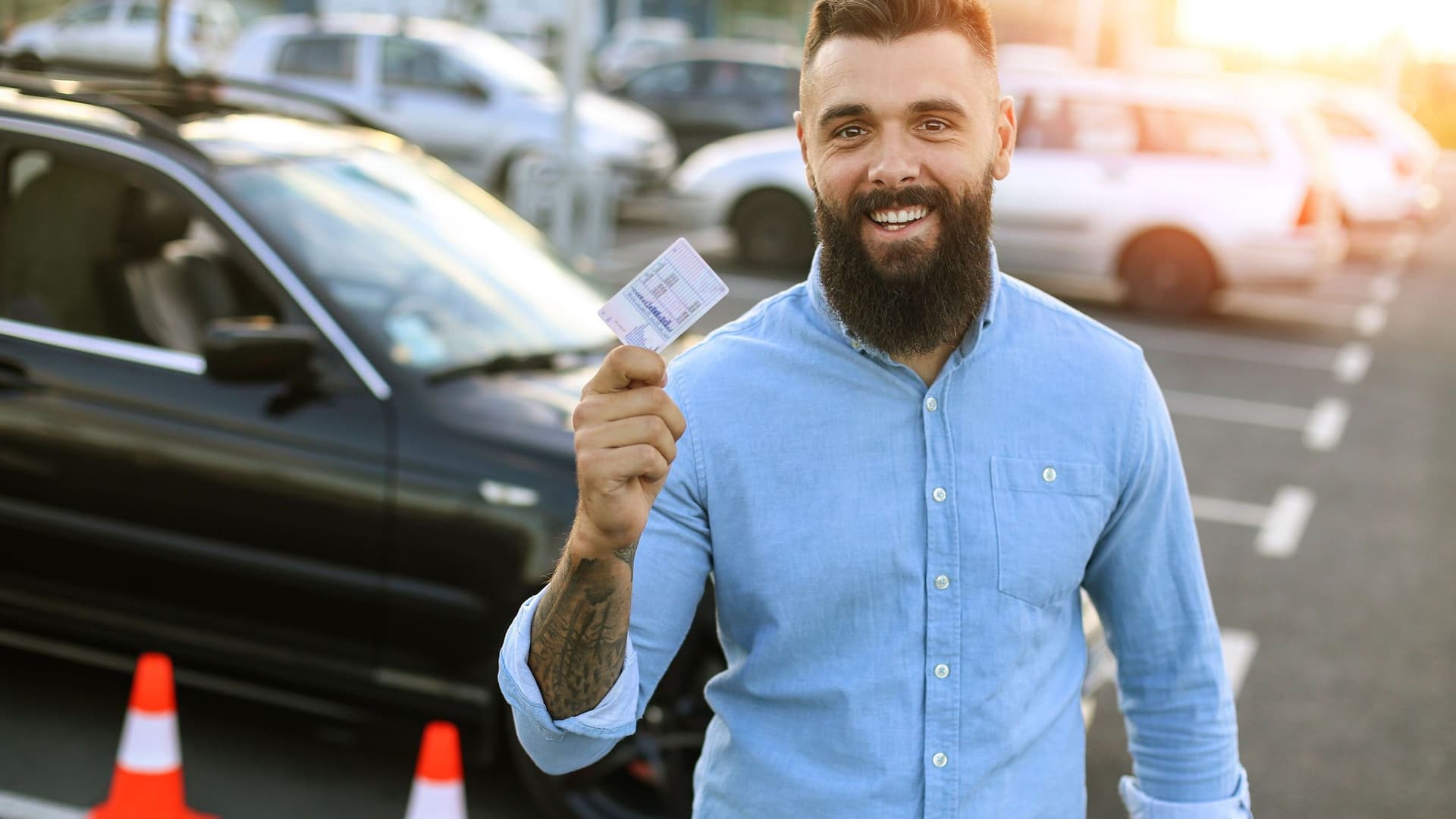 Junger Mann steht auf einem Parkplatz und hält lachend seinen Führerschein in die Kamera